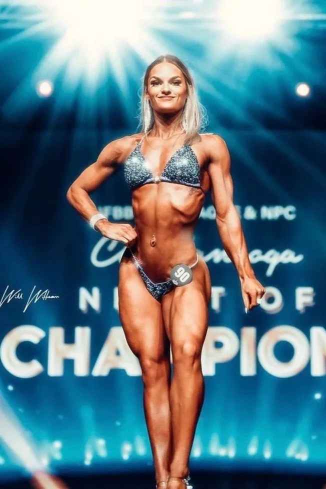 A female bodybuilder with long dark hair in a blue bikini and clear high heels posing on stage at a bodybuilding competition, with a bright blue background featuring the words "NPC CINCINNATI CHAMPIONSHIPS".