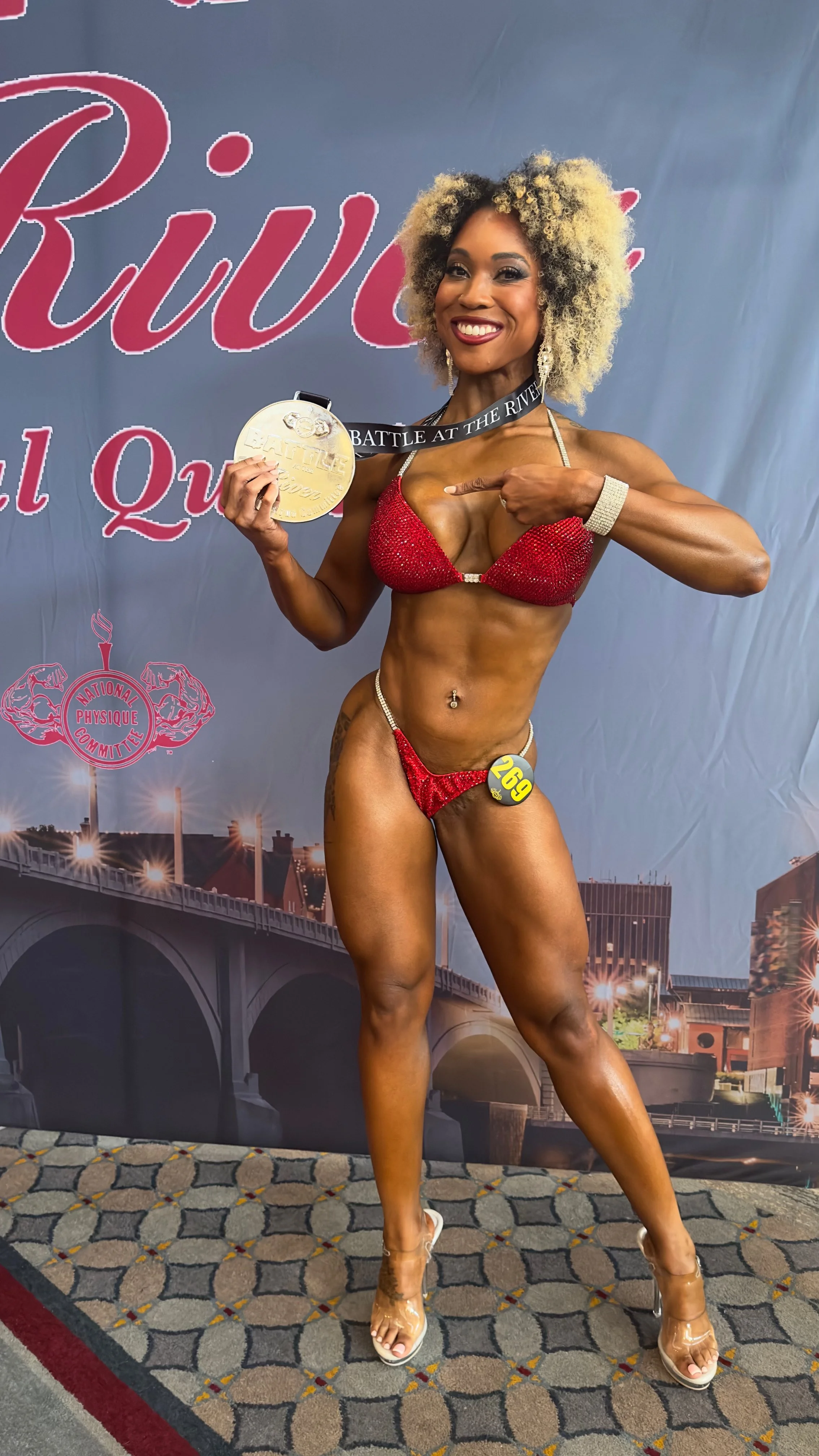 A female bodybuilder wearing a red bikini holding a gold medal, pointing at it with her other hand, standing on a patterned carpet in front of a backdrop with a city bridge and text.