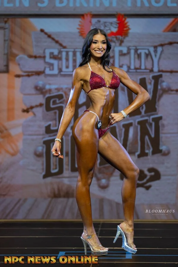 A female fitness competitor on stage during a bodybuilding competition, wearing a red sparkly bikini and clear high heels, posing with a smile against a backdrop with the words 'Sun City Bikini'.