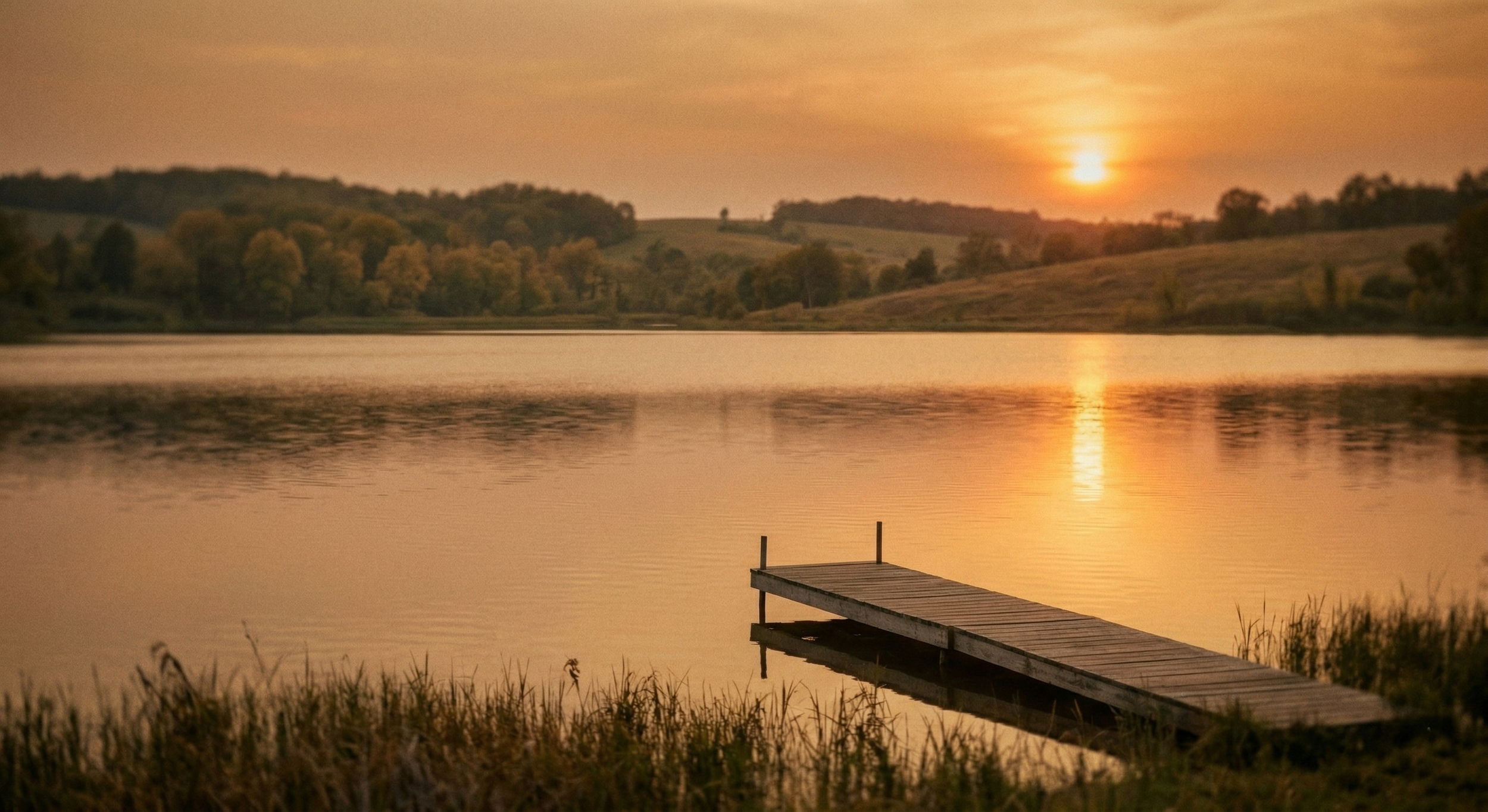 Sunset over a calm lake with a wooden dock in the foreground and rolling hills with trees in the background.
