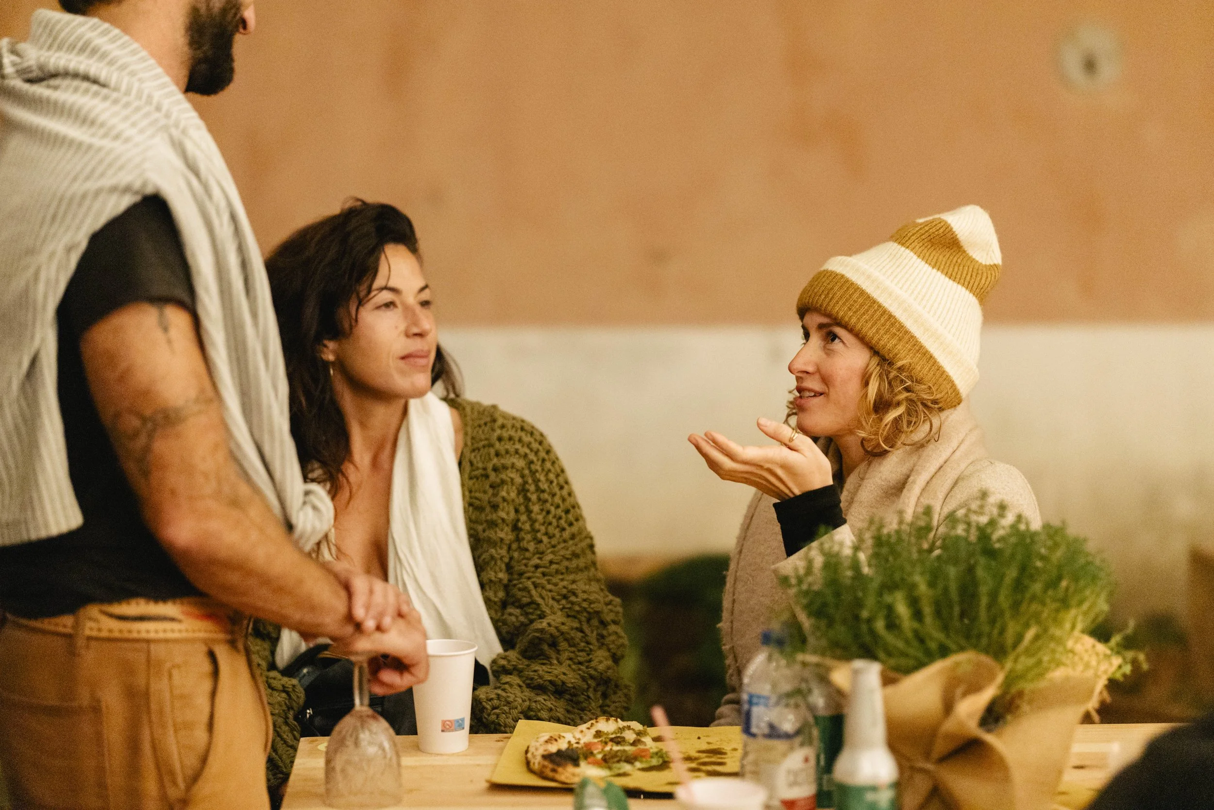Four people are gathered around a table, engaging in conversation. One woman is wearing a yellow and white striped beanie, and another woman is watching her. A man is standing on the side, and another woman holds a white cup. On the table, there is a pizza and some bottles and condiments, indicating a casual gathering or meal.