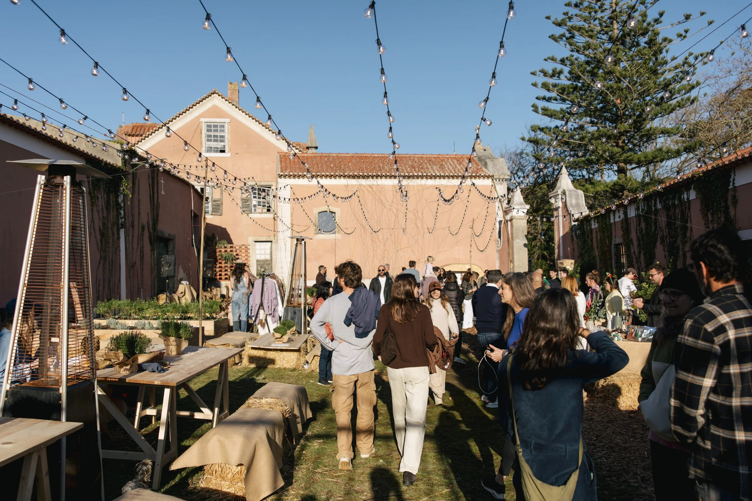 A lively outdoor market or festival scene with many people gathering and browsing various booths. String lights hang overhead, and there are trees and a brick building with a tiled roof in the background.