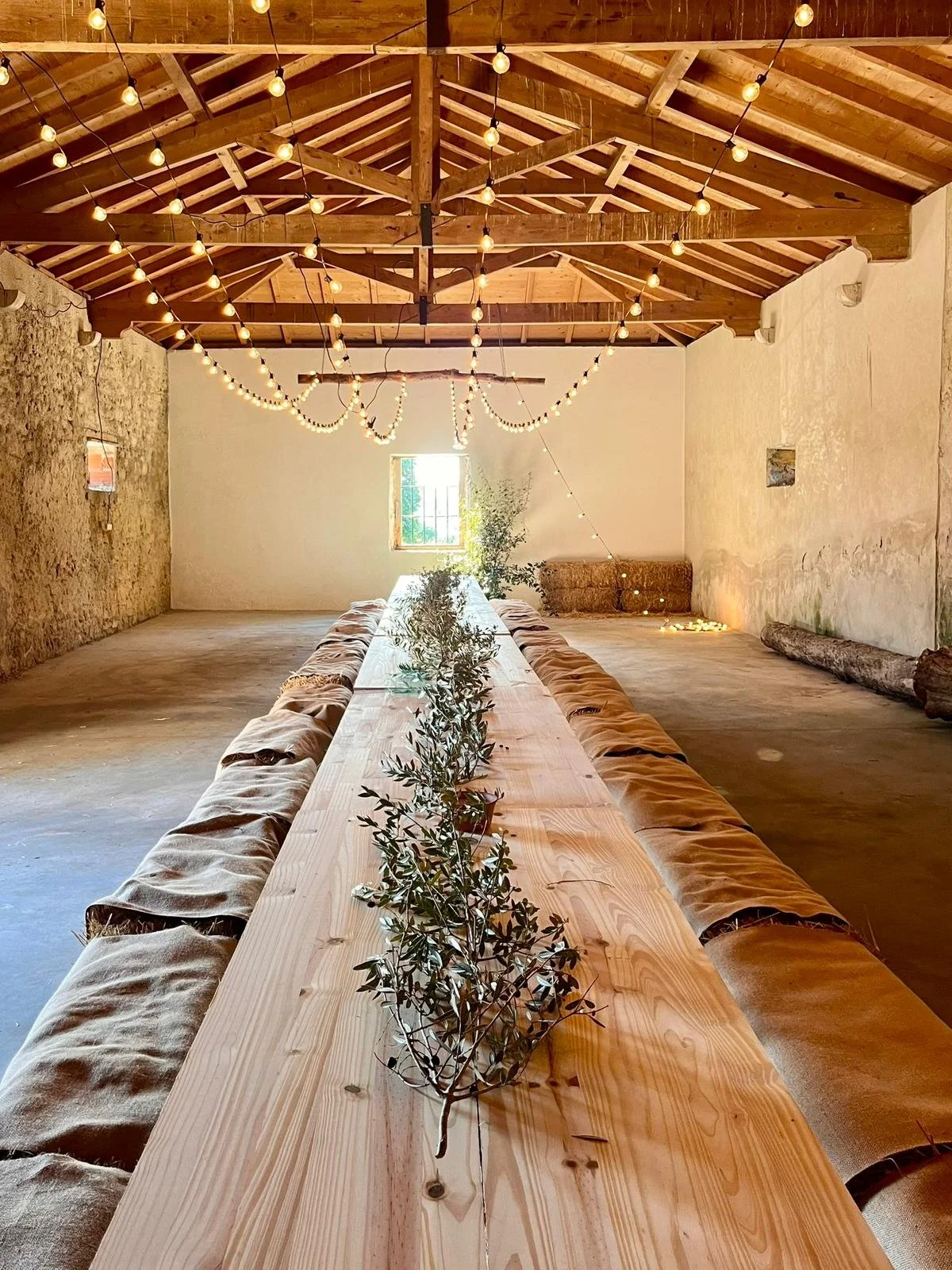 A rustic indoor event space with a long wooden table decorated with greenery, surrounded by cushioned benches. The ceiling features exposed wooden beams and string lights hanging overhead, creating a warm, cozy atmosphere.