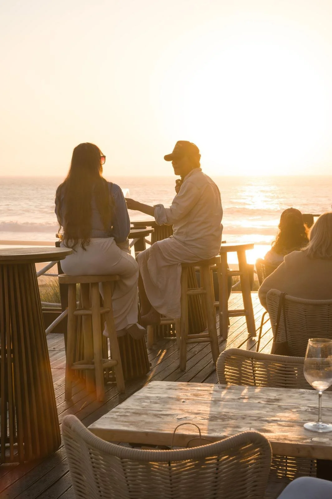 People sitting at an outdoor restaurant on a wooden deck by the ocean at sunset, engaging in conversation.