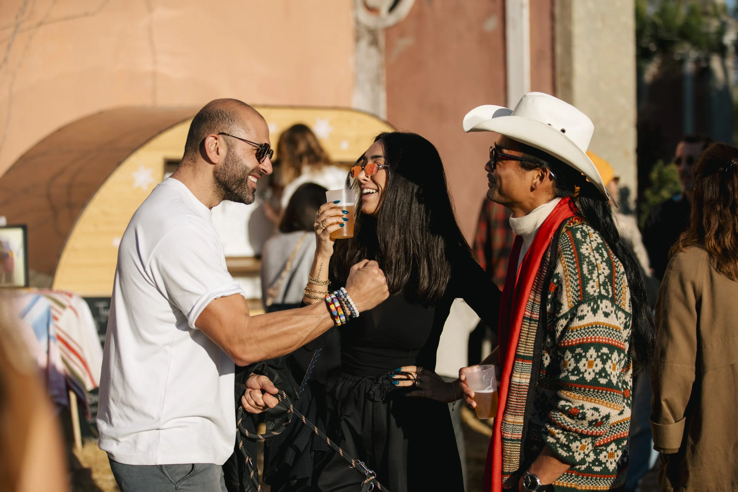 Three people are smiling and holding hands at an outdoor event. The man on the left has a beard, sunglasses, and is wearing a white shirt. The woman in the middle has long dark hair, sunglasses, and is holding a drink. The man on the right is wearing a white cowboy hat, sunglasses, and a colorful sweater. They seem to be enjoying themselves in a festive atmosphere.