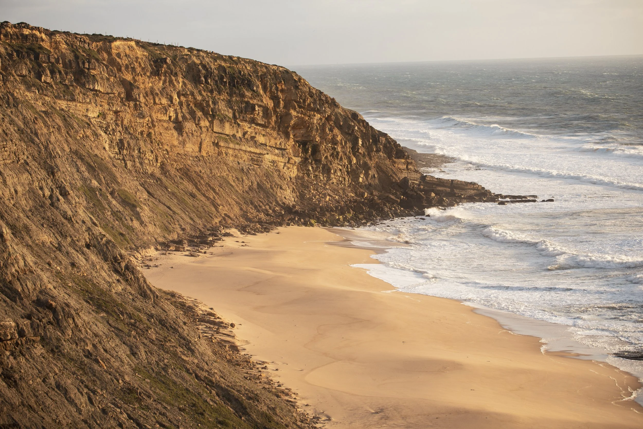 Cliffside overlooking the ocean with waves crashing onto the sandy beach below, under a partly cloudy sky during sunset.