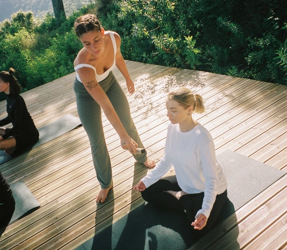 Two women practicing yoga on a wooden deck outdoors; one is sitting cross-legged in a meditative pose, and the other is standing and assisting.