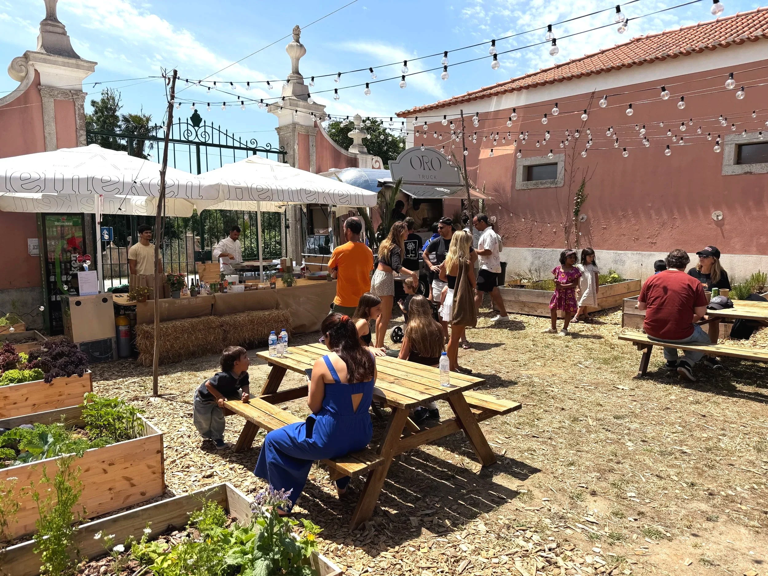 Outdoor gathering with people socializing and sitting at picnic tables under string lights, a pink building with small windows in the background, and vegetable garden beds in the foreground.