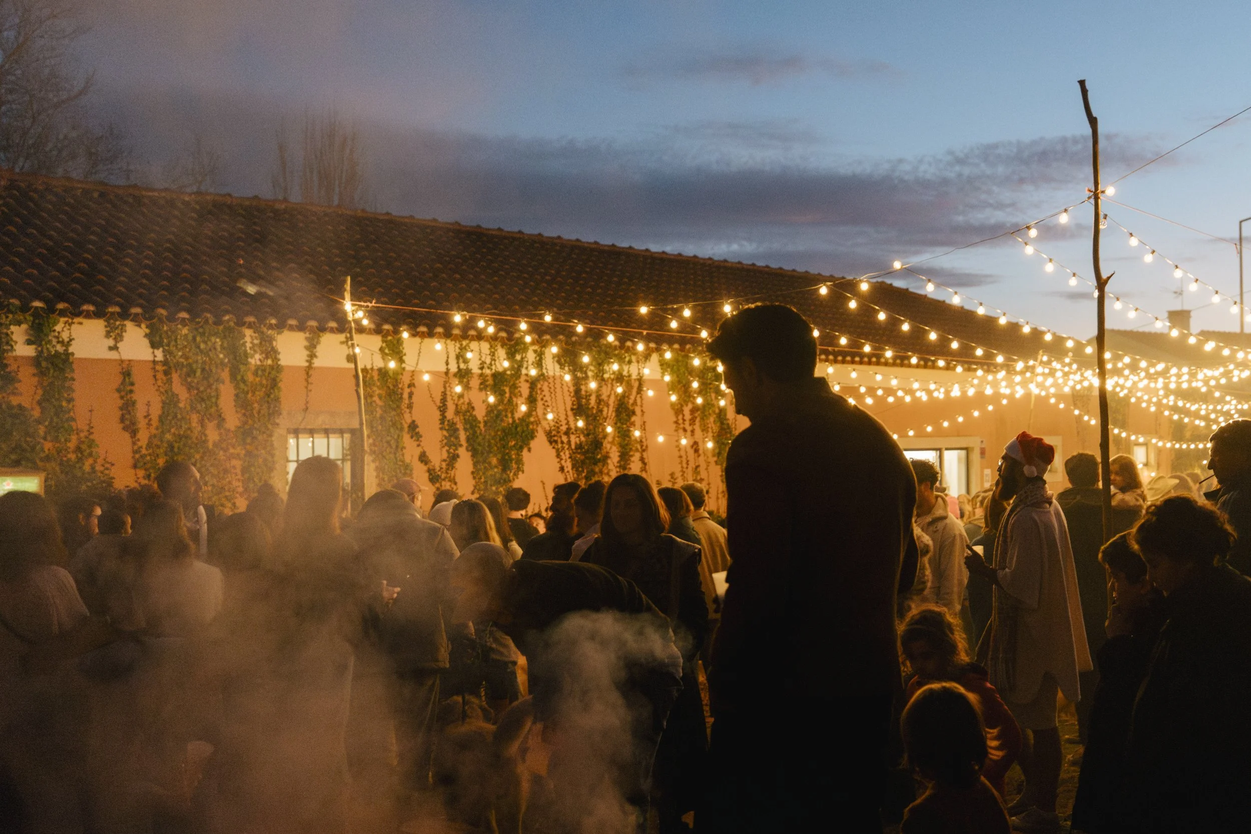 Crowd of people gathered outdoors during evening, with string lights hanging above and a building with a tiled roof in the background.