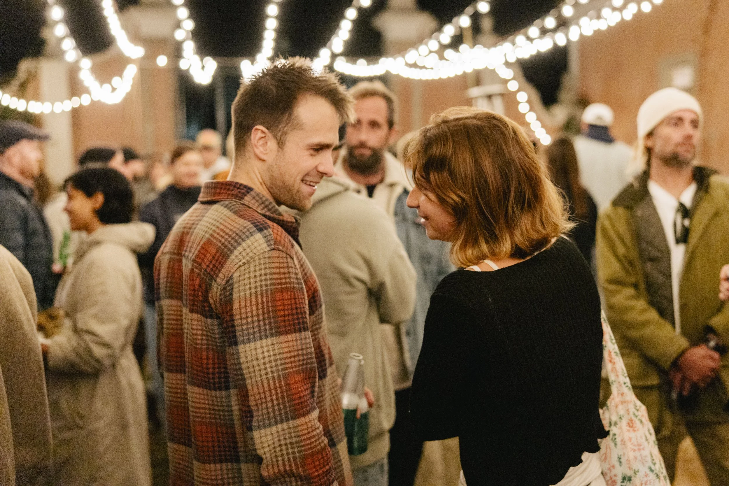 A young man and woman are smiling at each other at a social gathering under string lights, with other people mingling in the background.