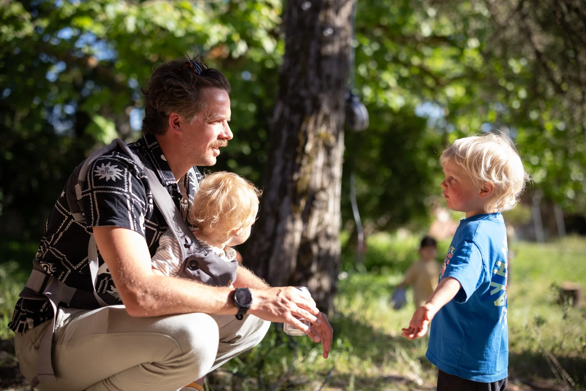 A man talking to a young boy outdoors in a park with trees in the background.