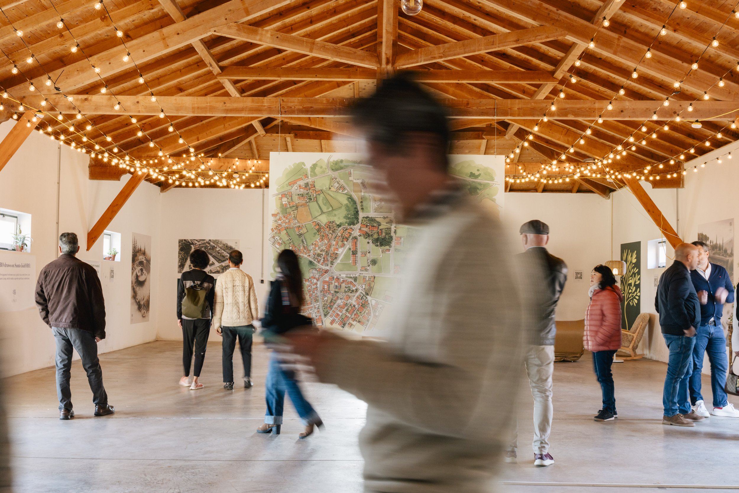People walking and standing in an exhibition hall with wooden beams and string lights, viewing maps and artwork on the walls.