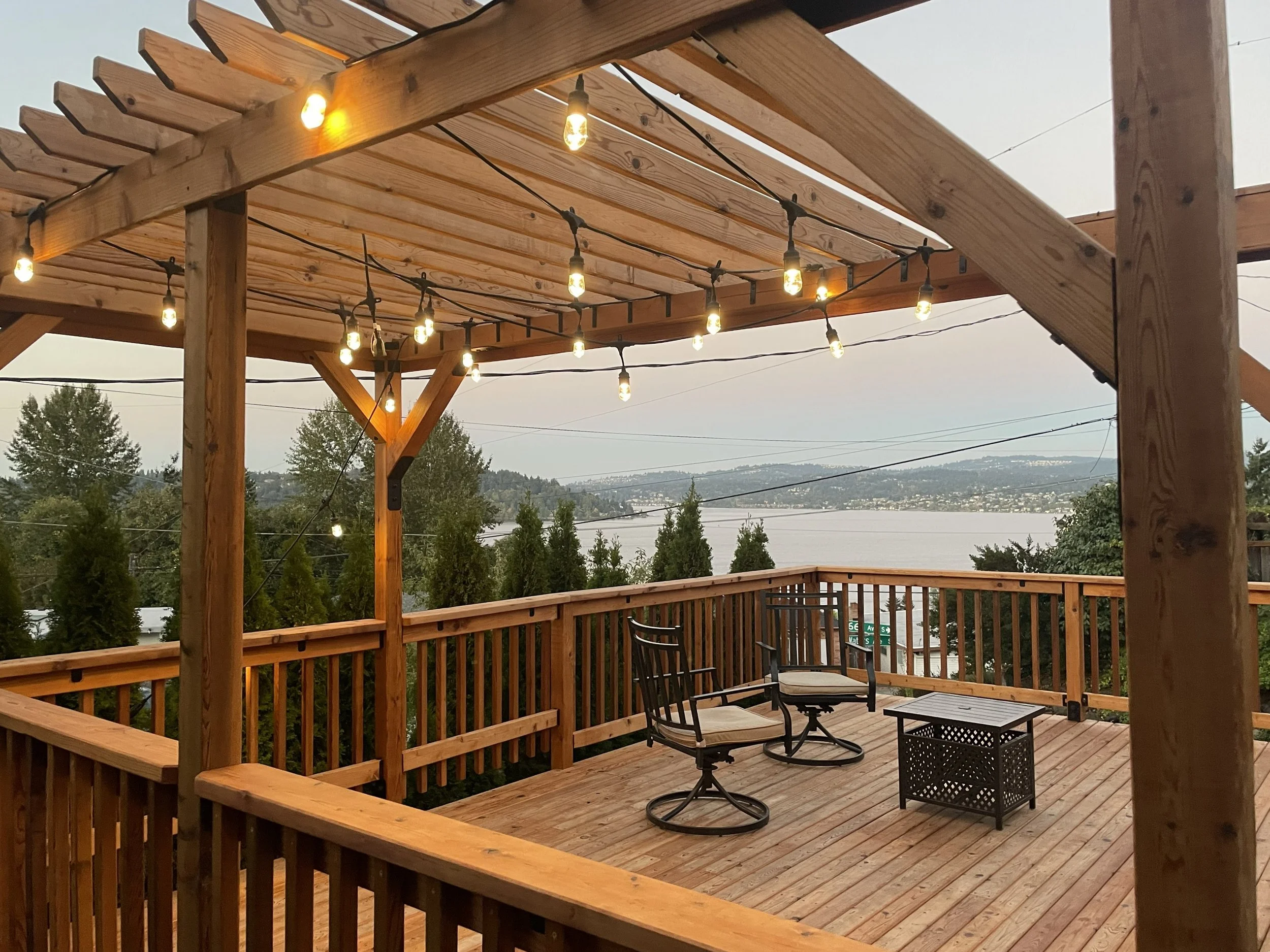 Wooden outdoor deck with string lights, two metal chairs with cushions, a small black table, overlooking water and trees with hills in the distance.
