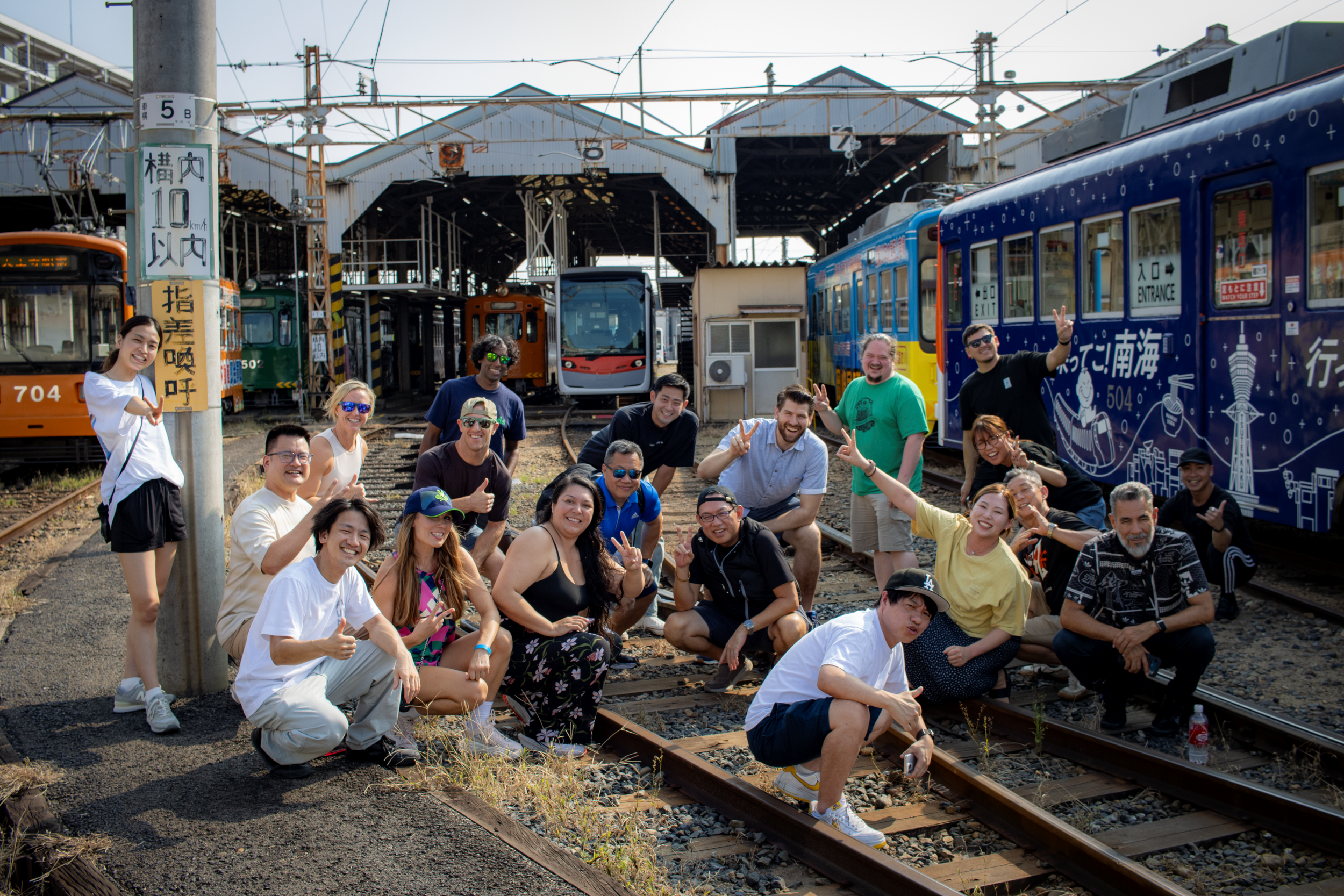 A group of people smiling and posing on railroad tracks in front of various colorful trains and train stations.