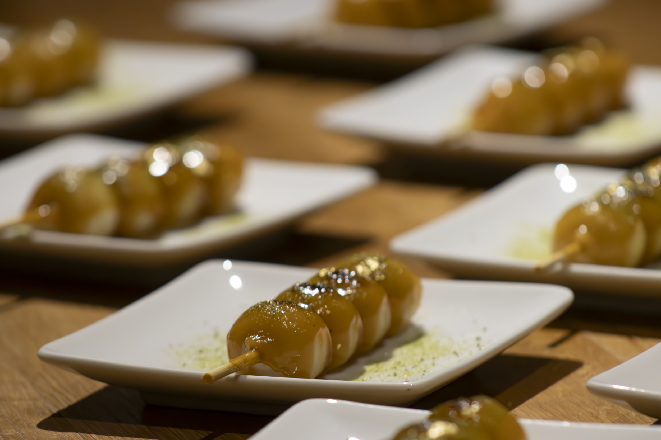 Skewers of grilled meat with sauce on white plates arranged in a row on a wooden table.