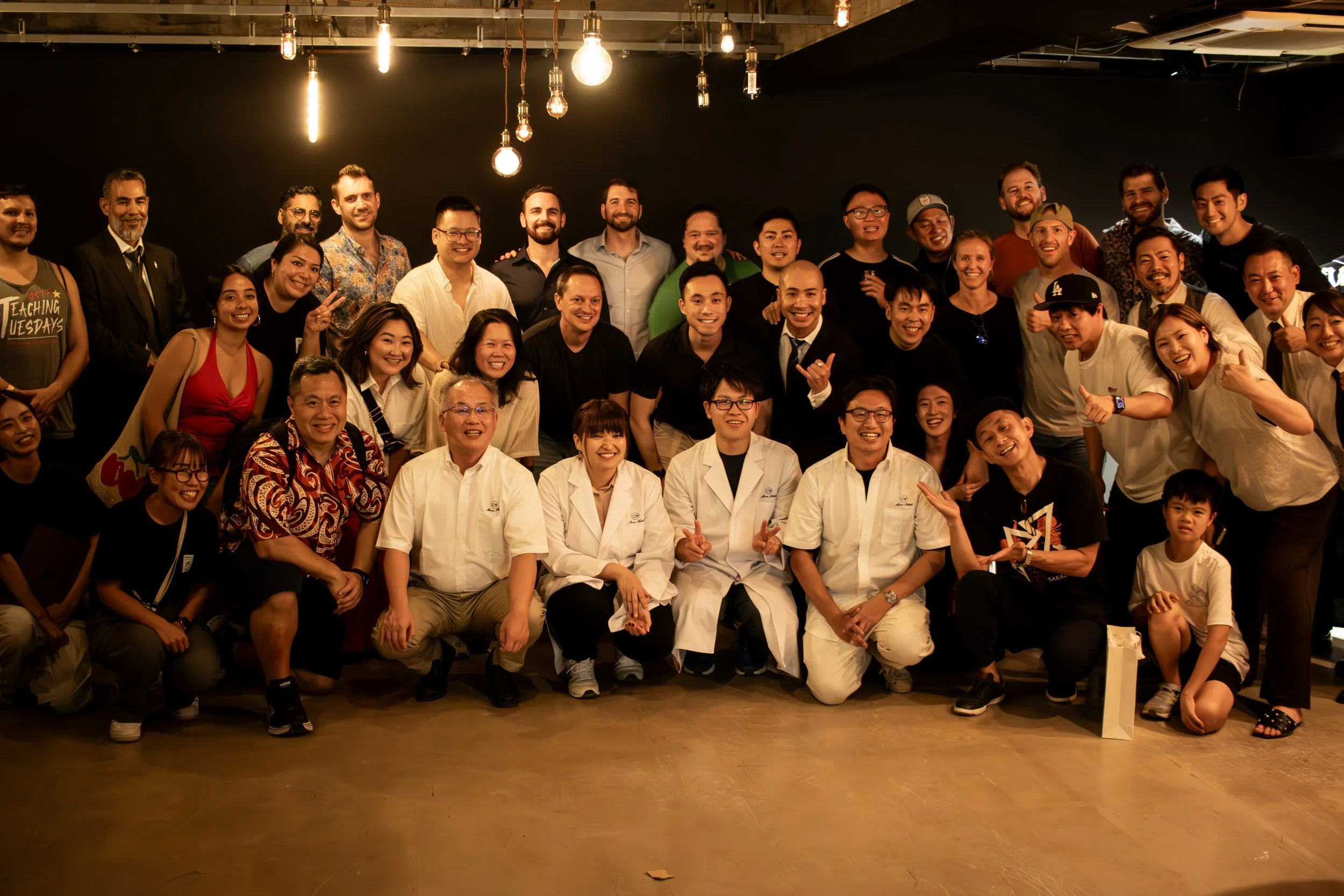 Group of people posing for a photo indoors under hanging light bulbs, smiling and making gestures, with some dressed in white coats.