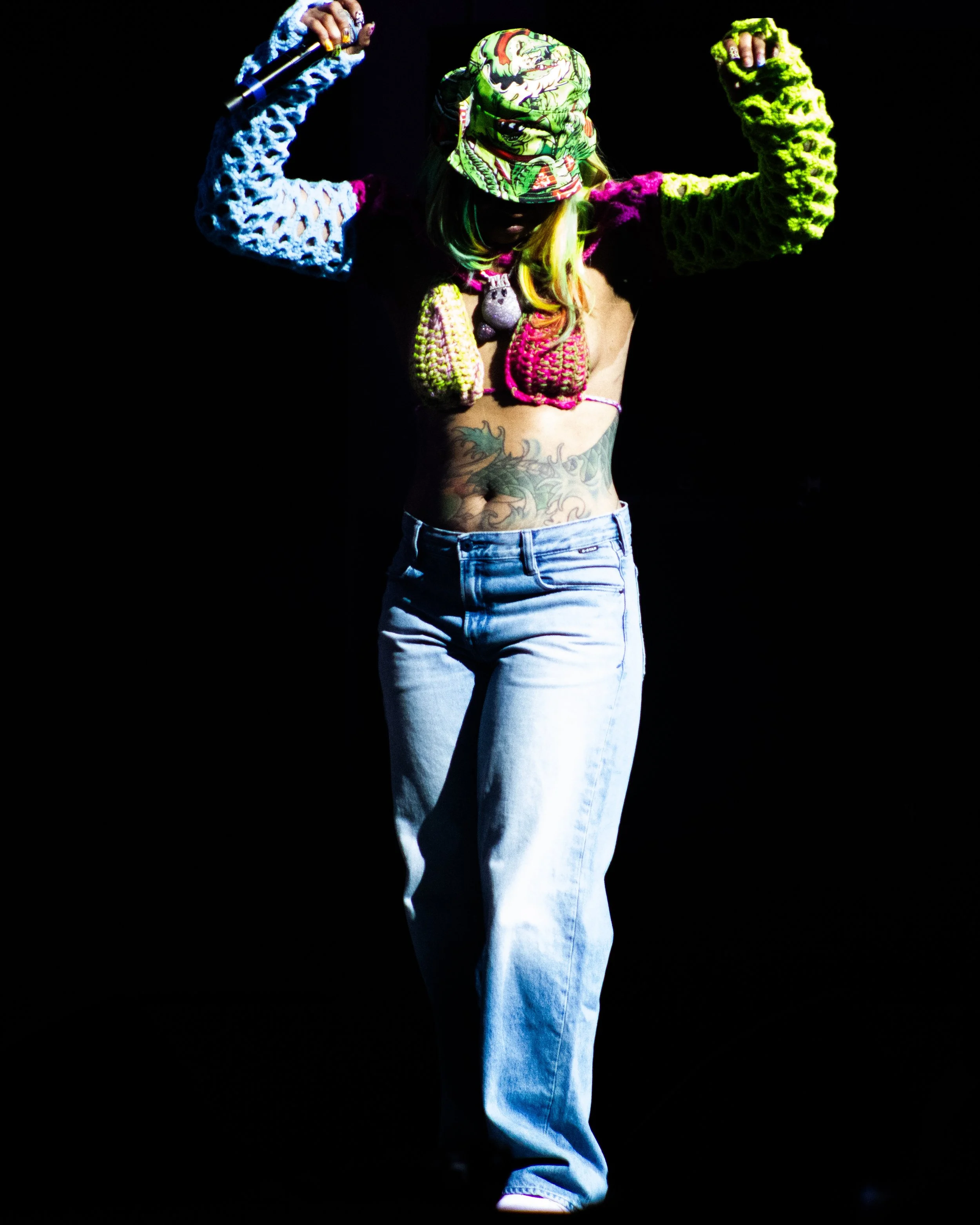 Person wearing a colorful bucket hat, multicolored crochet jewelry, and loose blue jeans, holding a microphone with arms raised in a dramatic pose against a dark background.