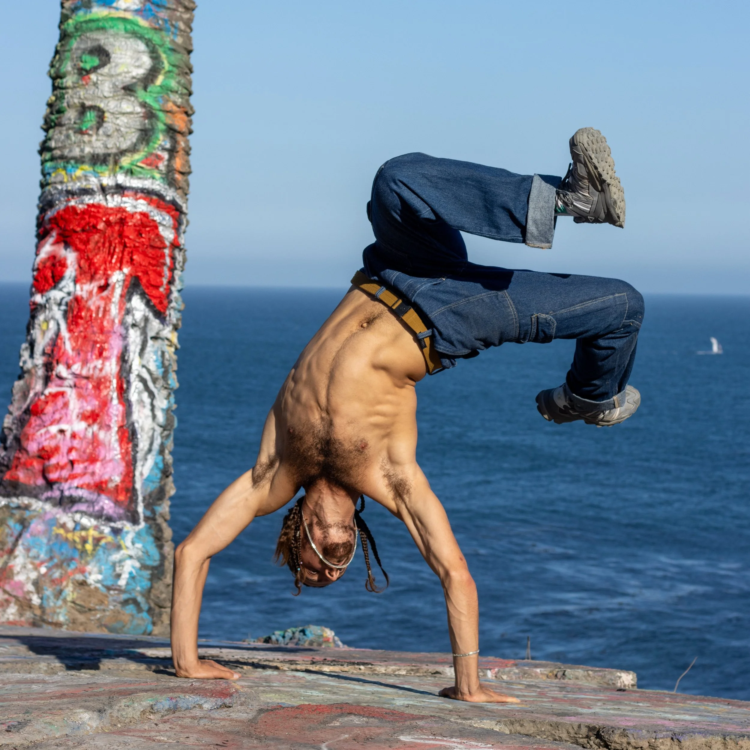A woman is performing a handstand on a colorful, graffiti-covered platform by the ocean, with her legs bent at the knees and feet pointing sideways.