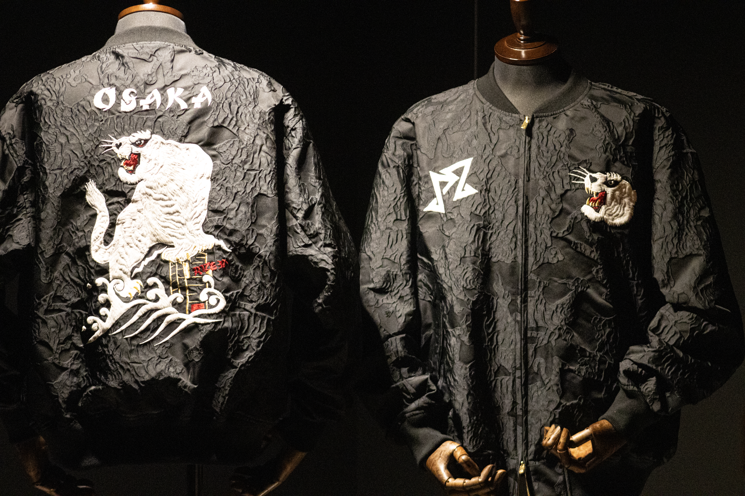 Mannequin dressed in a black bomber jacket with textured fabric, embroidered white tiger on the back, and white logo and symbols on the front and back.