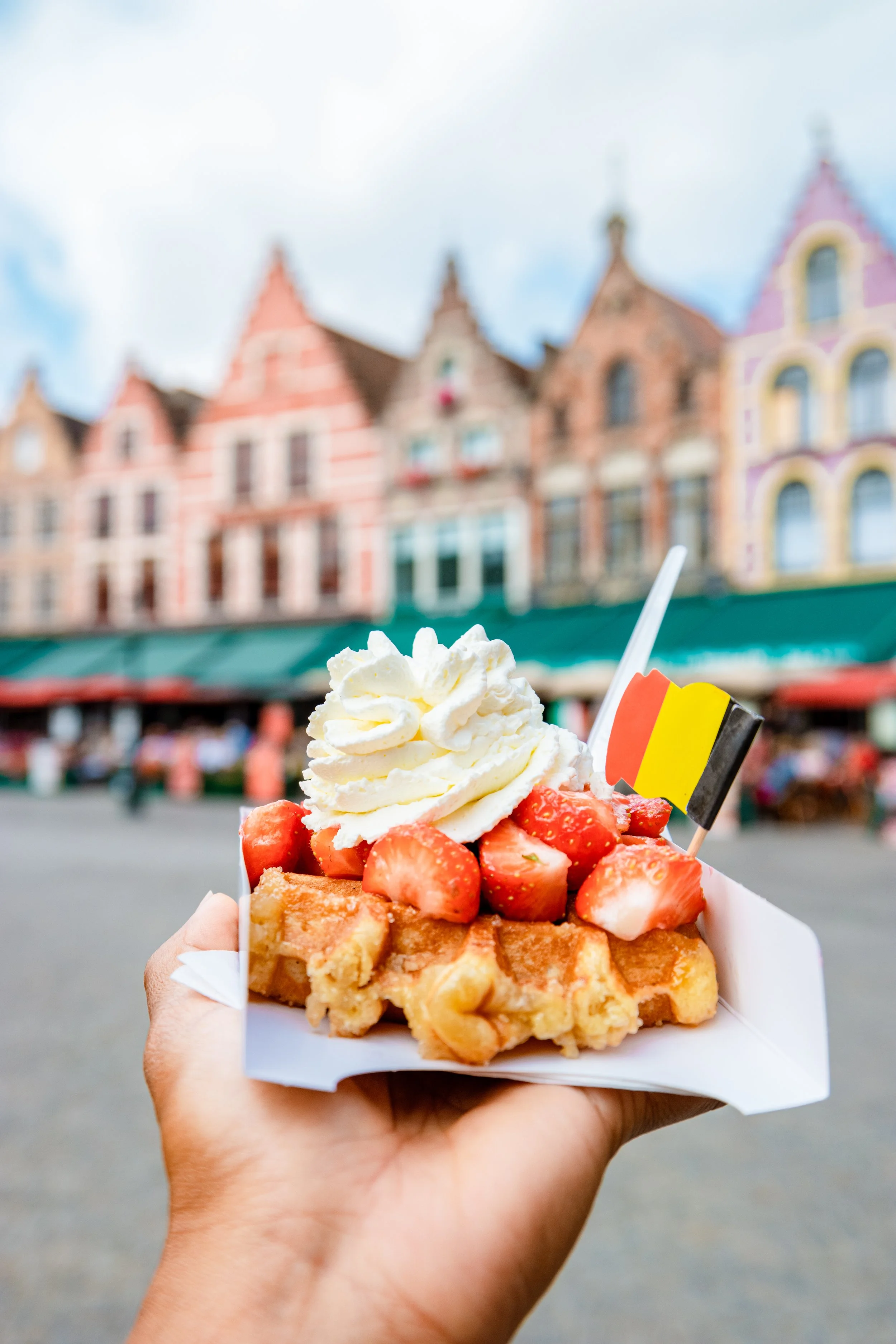 Hand holding a waffle topped with strawberries, whipped cream, and a small Belgian flag against a colorful European-style town square.