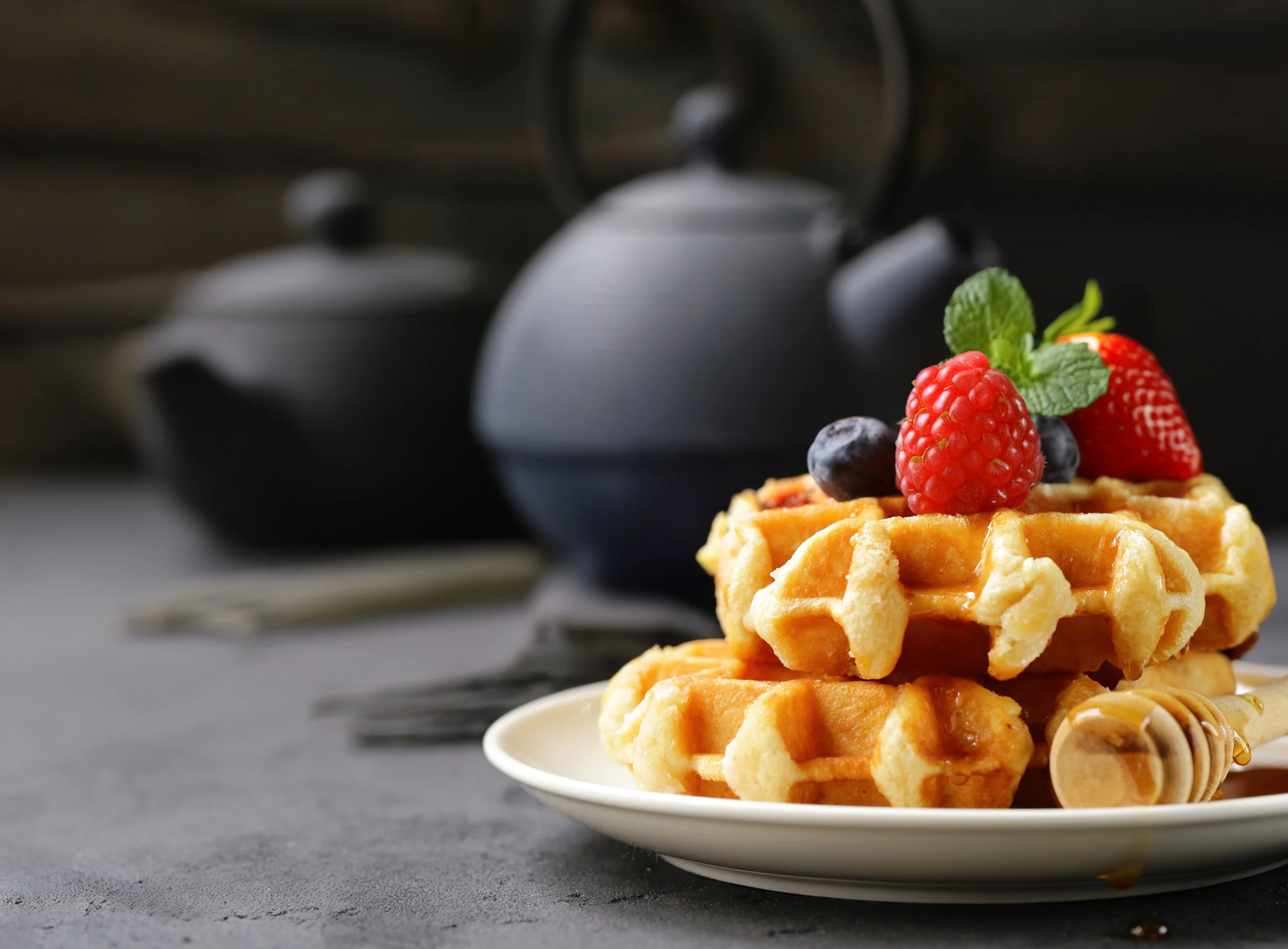 Waffles topped with strawberries, blueberries, and mint leaves on a white plate, with a black teapot in the background.