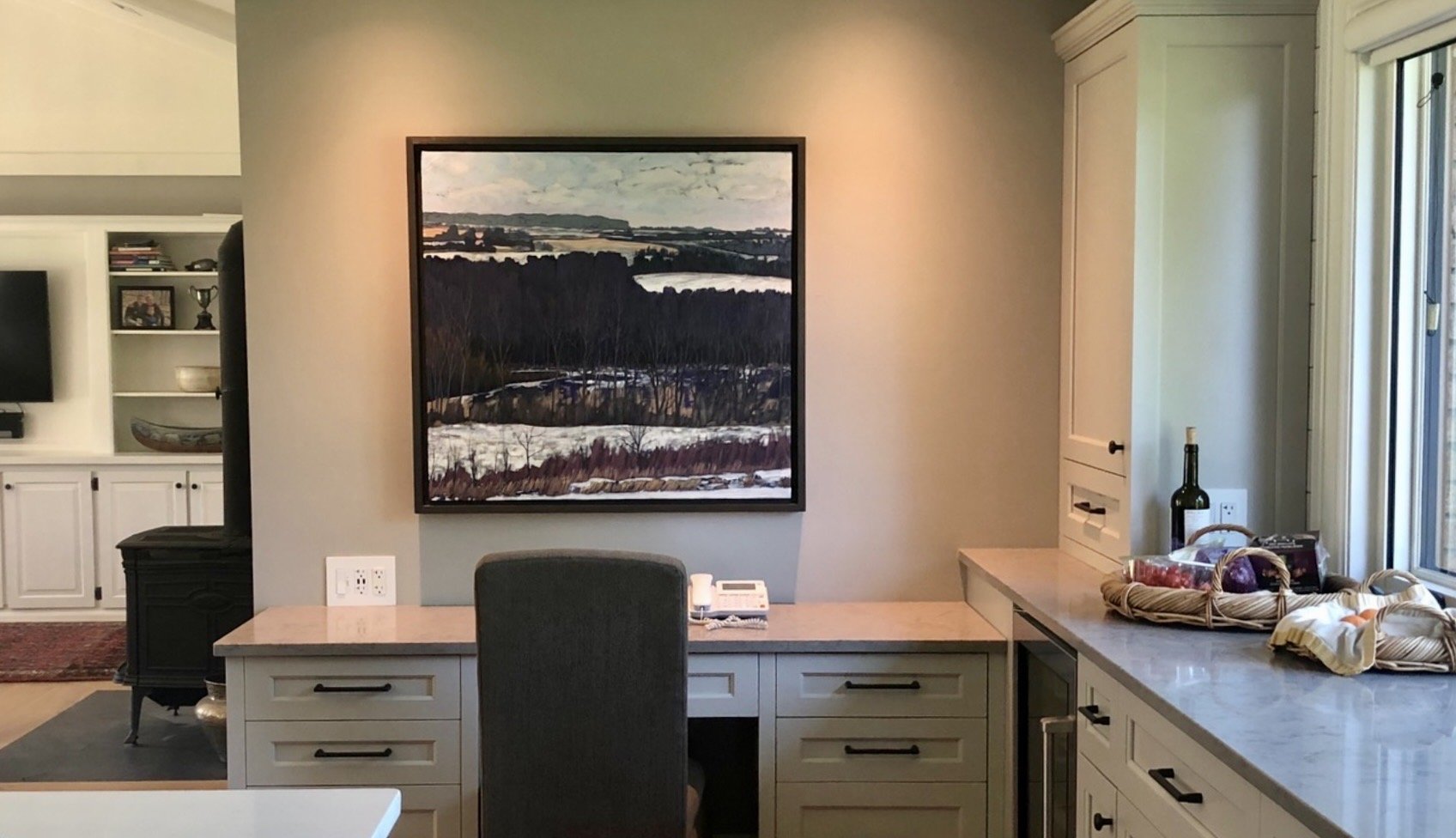 Living room with a desk, framed landscape painting, window with sunlight, and kitchen counter with baskets and a bottle.