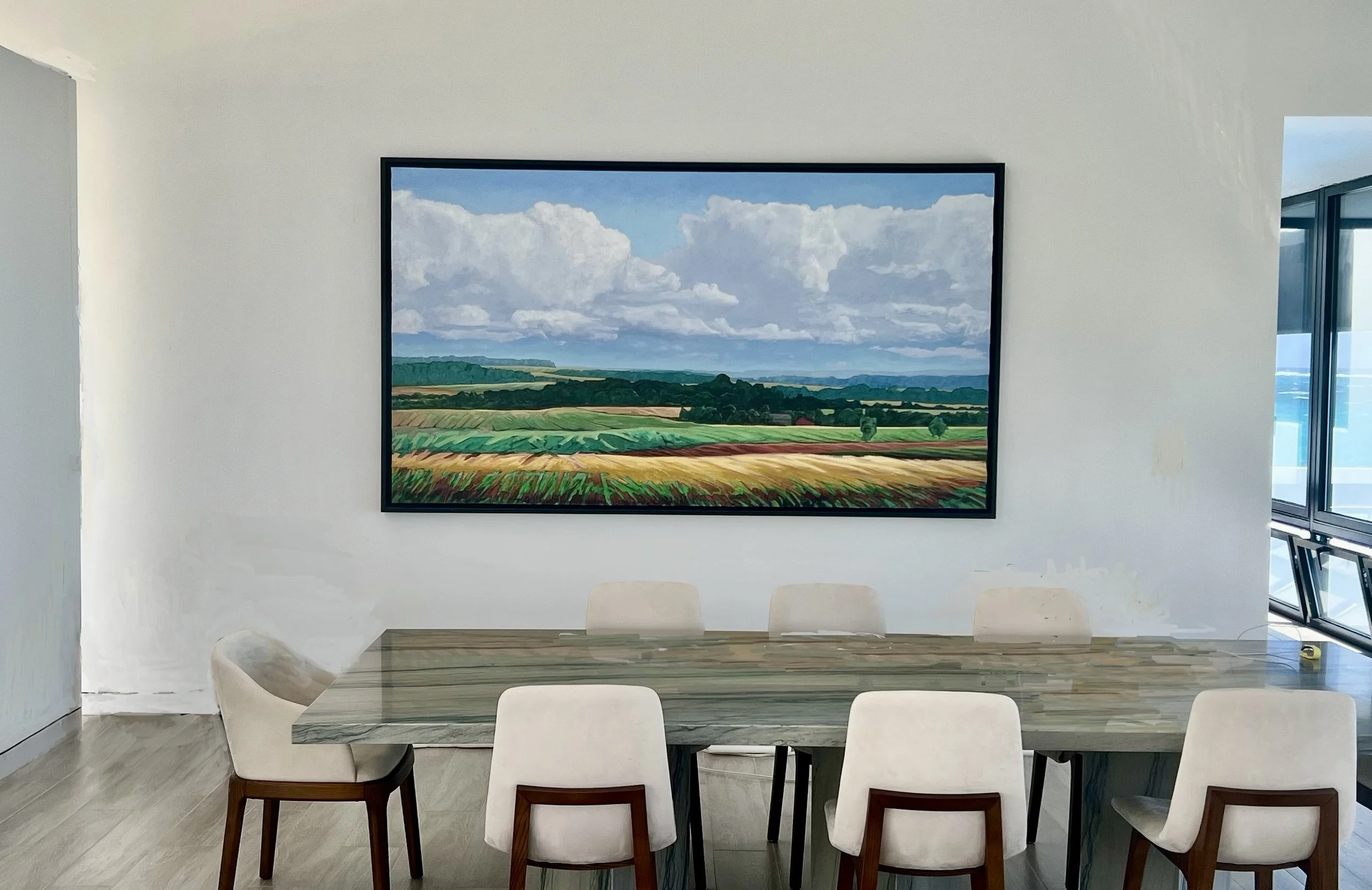 Contemporary dining room with a large landscape painting of fields and sky on the white wall, six beige chairs around a rectangular wooden table, and a window with a view of the building exterior.