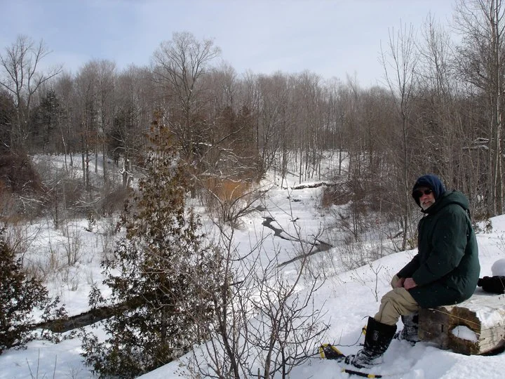 A man sitting on a log in a snow-covered landscape with trees, a small creek, and a cloudy sky.