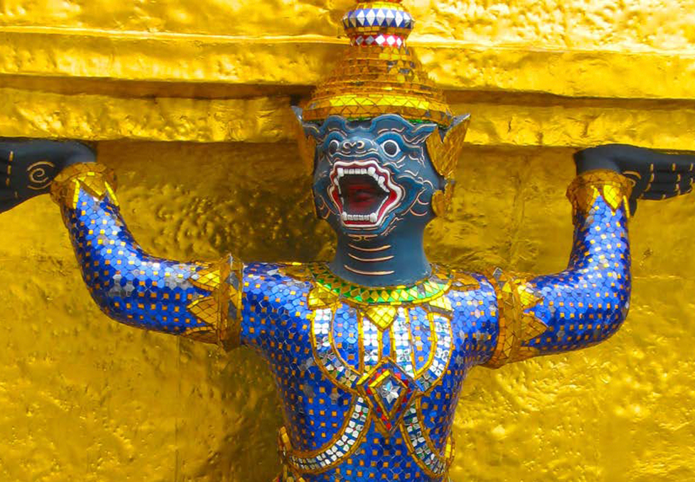 A colorful, mosaic statue of a muscular, mythical creature with a blue face, open mouth, and golden, ornate body, holding a golden horizontal bar against a textured golden background.