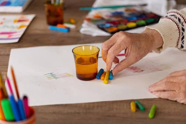 Person's hand placing small colored chalks on a white paper next to a glass of brown liquid on a wooden table with art supplies in the background.