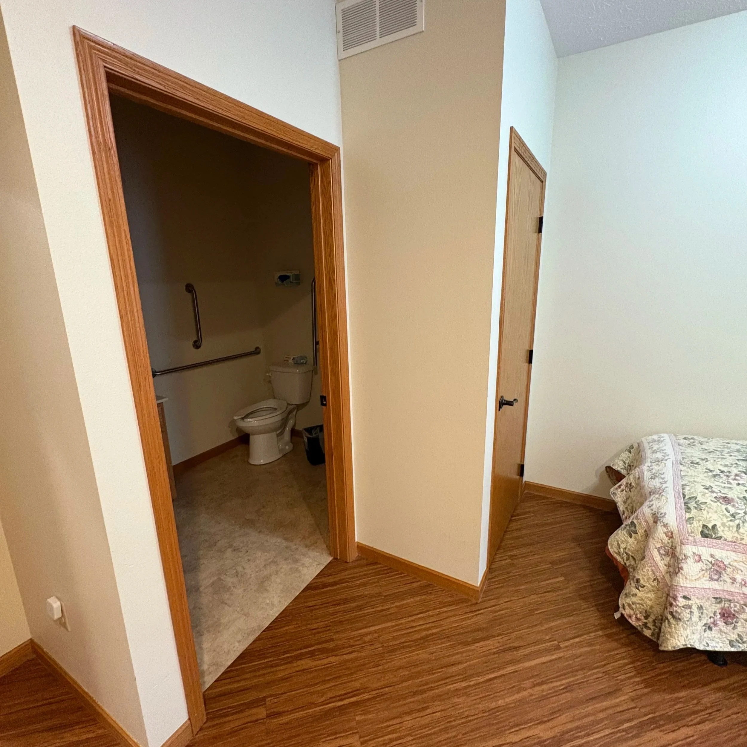 View of a bedroom corner with an open doorway leading to a bathroom. The doorway has a wooden frame, and the bathroom contains a toilet. The room has light-colored walls, wood flooring, and part of a floral-patterned bedspread is visible on a bed in the lower right corner.