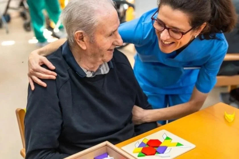 A woman in blue scrubs and glasses smiling with a elderly man in a dark sweater, sitting at a table with a board game and laughing together.