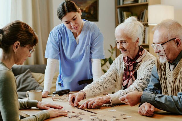 A healthcare worker and three elderly people working on a puzzle at a dining table.