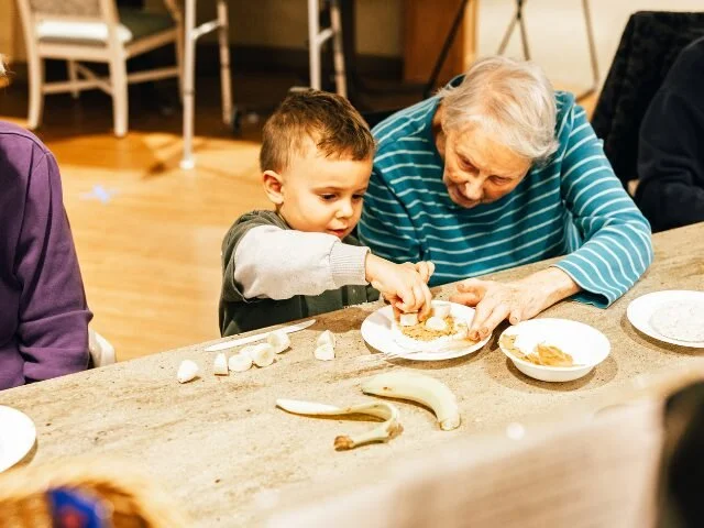 An elderly woman and a young boy sitting at a kitchen counter, sharing breakfast with torn pieces of bread and a banana on the table.
