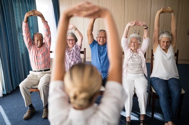 A group of senior people sitting on chairs and participating in a seated exercise or stretching class, with a woman leading in the foreground.