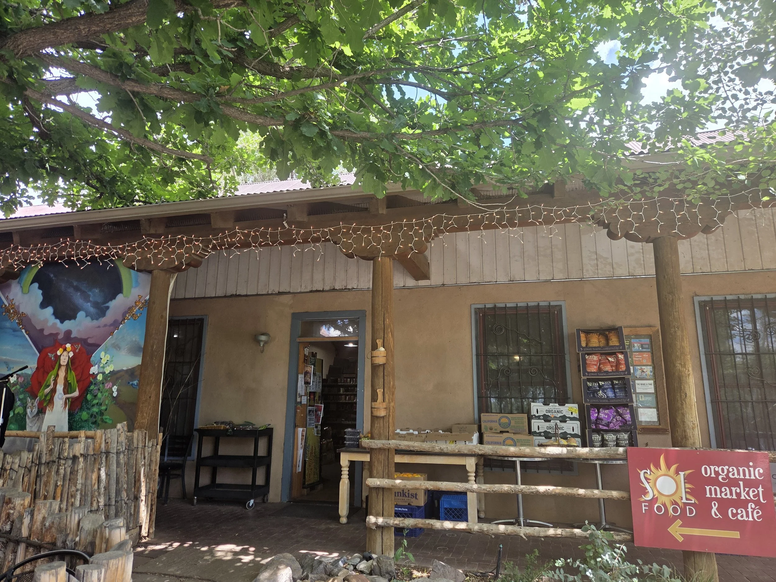The image shows an outdoor view of a building with a wood and metal exterior, decorated with string lights under a large tree with green leaves. There is a colorful mural of a woman on the left. A sign on a red stand indicates a natural food and cafe market.