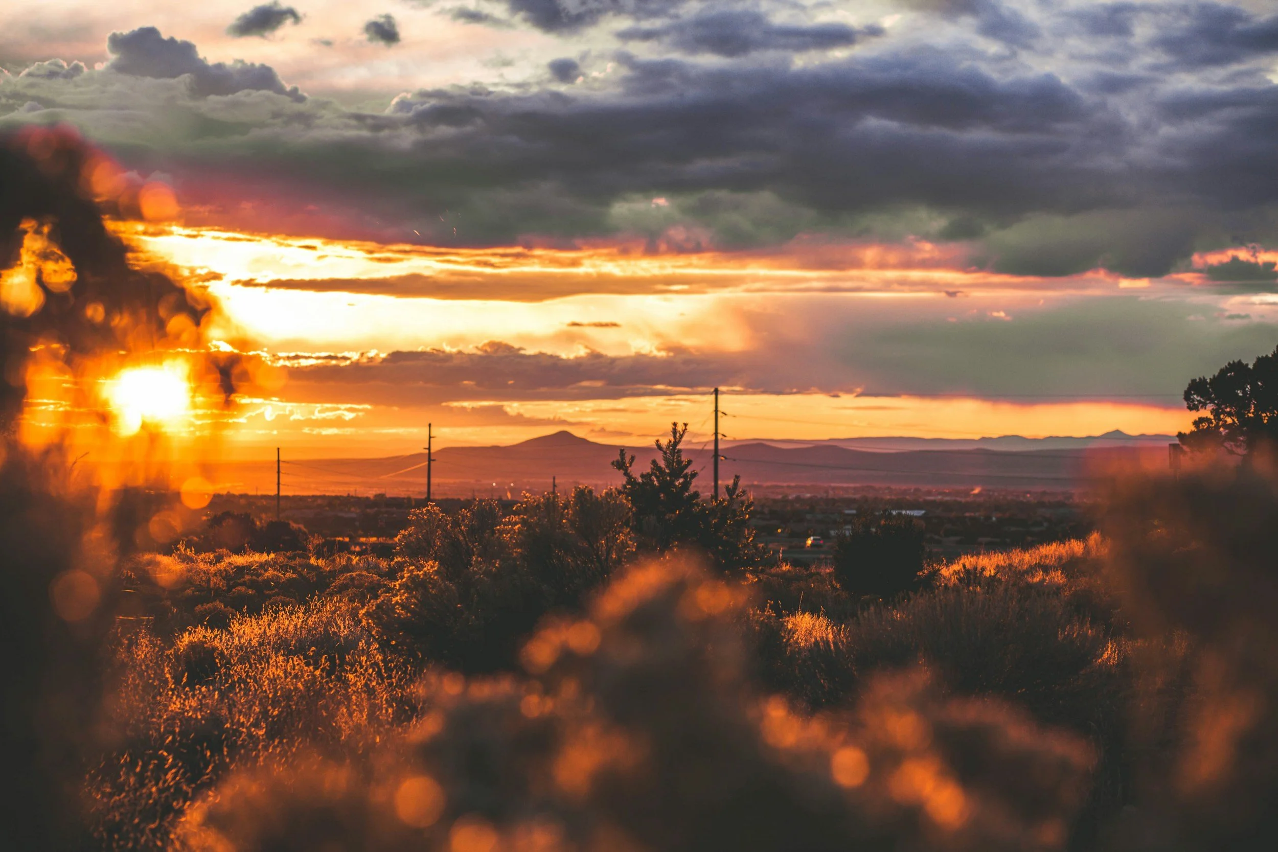A vibrant sunset over a rural landscape with dark, cloudy skies, mountains in the background, and power lines in the foreground.
