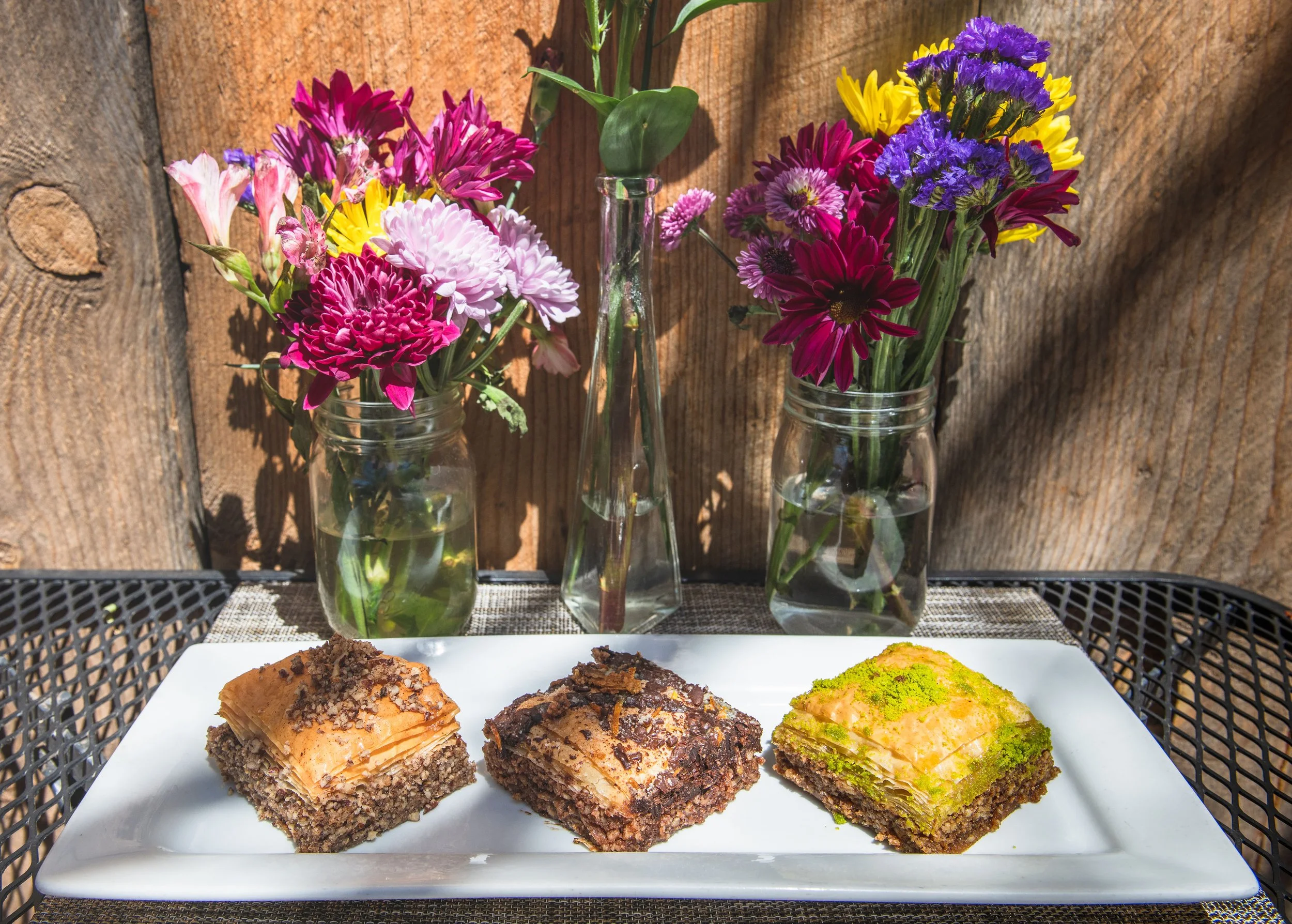 A white rectangular plate with three pieces of layered cake topped with chocolate crumbles and green powder, placed on a black metal outdoor table with a wooden wall and three glass bottles with colorful flowers in the background.