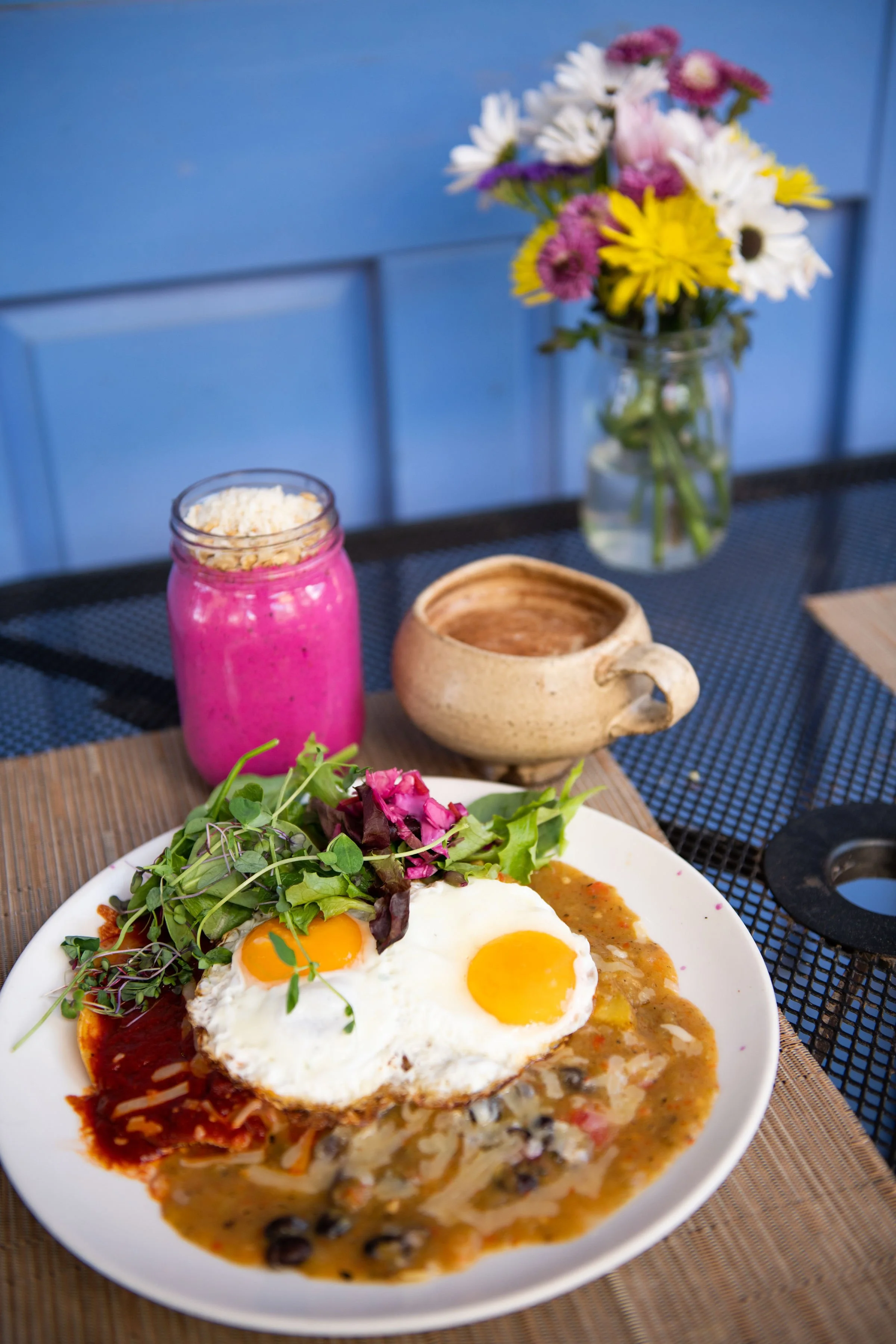 A plate of breakfast food including eggs, beans, and salsa, topped with microgreens, served on a table. In the background, there is a vase of colorful flowers and a purple smoothie in a jar. The scene is set on a dining table with a placemat and drinks.