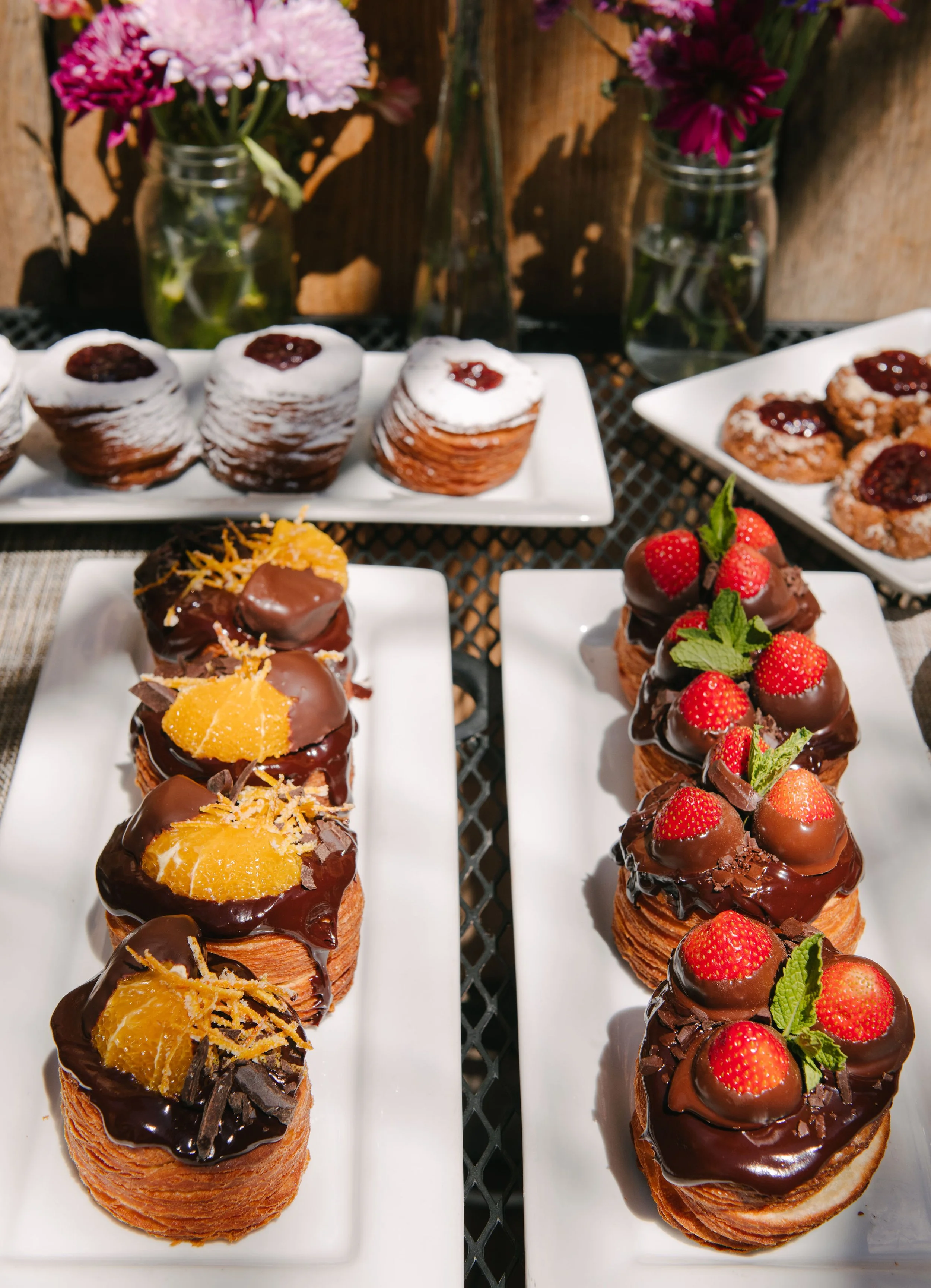 Assorted pastries topped with chocolate, fruit, and garnishes, displayed on white plates with flower vases in the background.