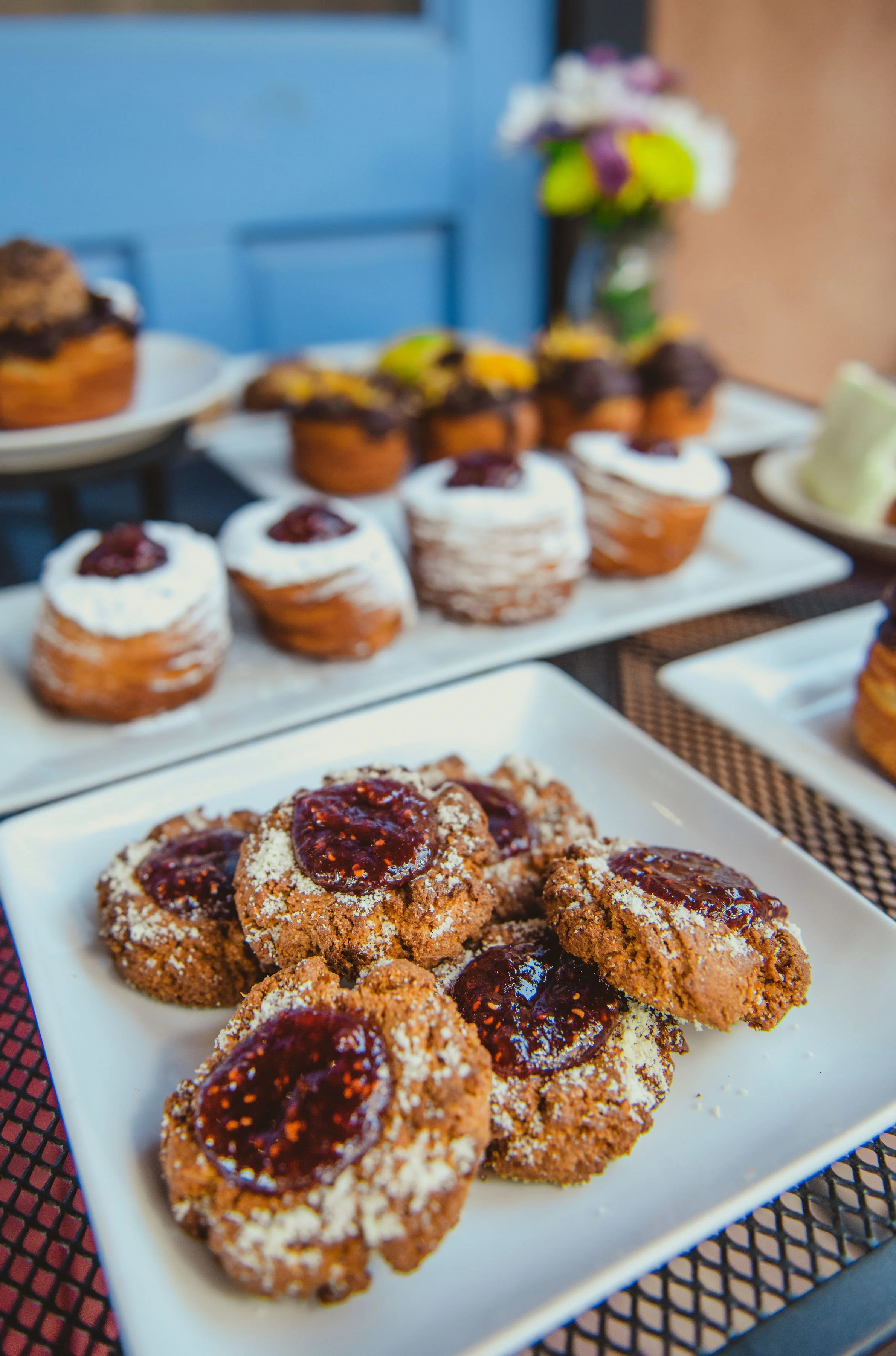 Close-up of cookies with jelly on a white rectangular plate, with blurred cupcakes and a flower vase in the background.