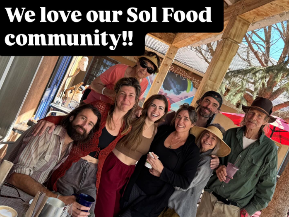 Group of eight people gathered outdoors under a wooden structure, smiling and enjoying each other's company, with food and drinks on the table. A banner reads 'We love our Sol Food community!!'.