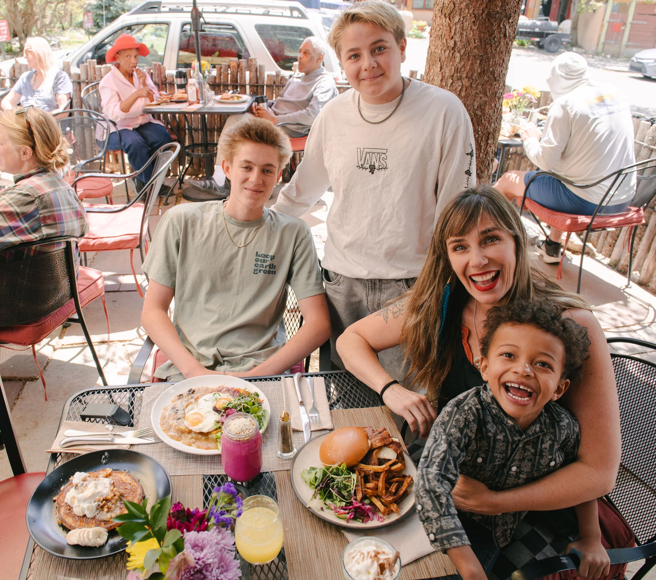 A group of people enjoying an outdoor meal at a restaurant table, with several dishes including pizza, fries, and drinks, under a large tree with background of other diners, a white car, and a street.