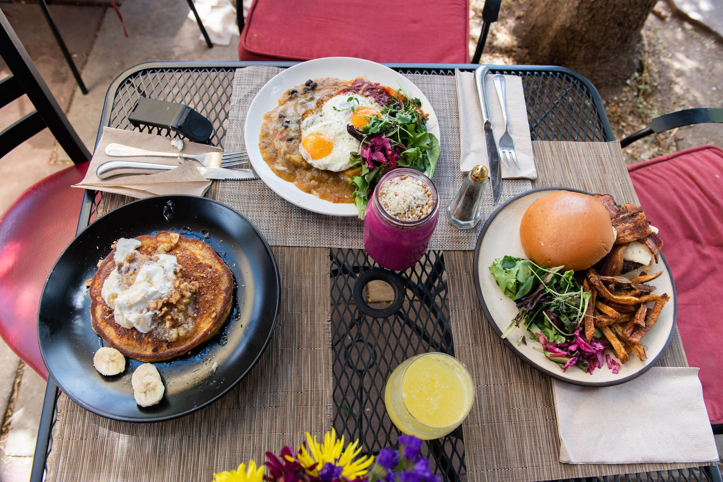 Outdoor breakfast table with plates of food including pancakes with whipped cream, eggs, salad, a burger with fries, a purple and pink drink, and a yellow drink, with flowers in the foreground.