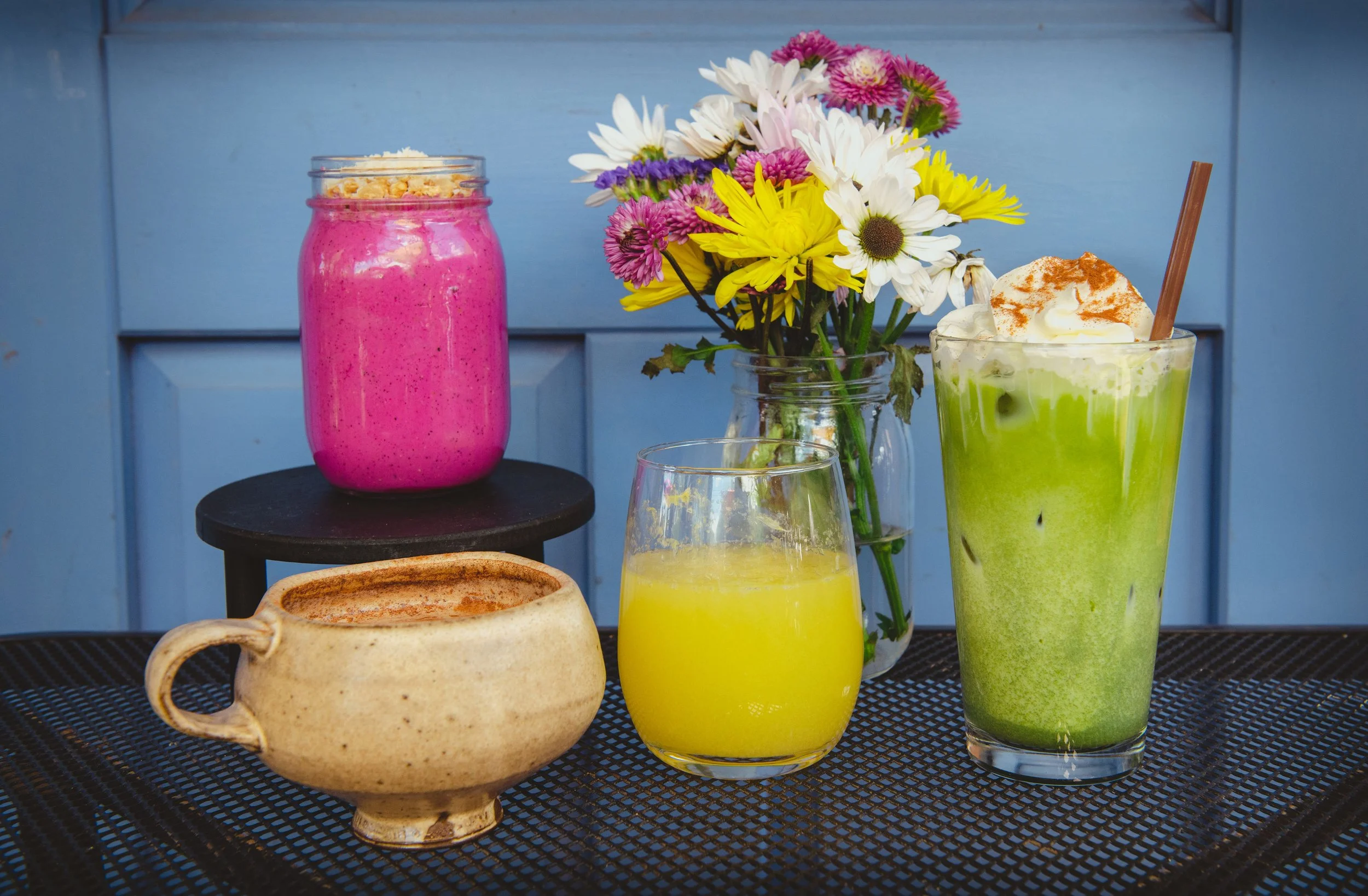 A table with a variety of colorful beverages, a bouquet of flowers, and a ceramic mug on a dark surface against a light blue wooden background.