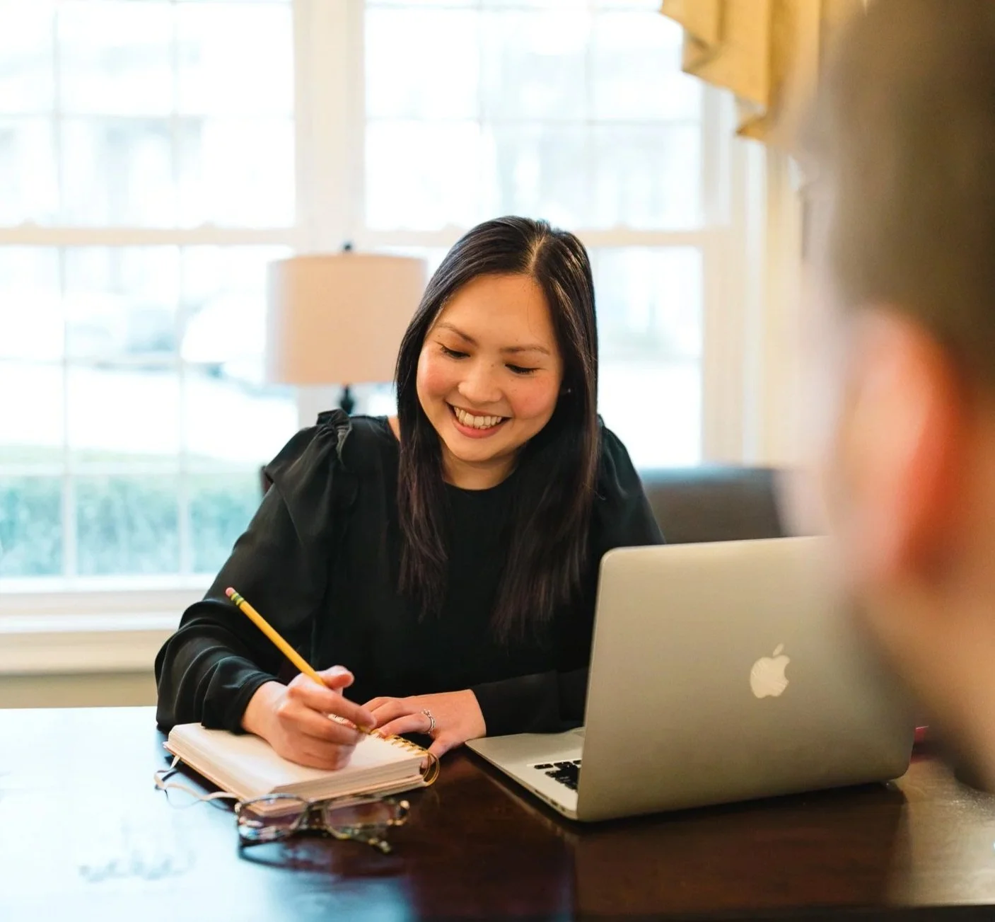A young woman with dark hair smiling while writing in a notebook, sitting at a table with a silver MacBook laptop in front of her, in a brightly lit room with large windows. Bing Gonzalez Coaching. Professional Neuroscience Coach.