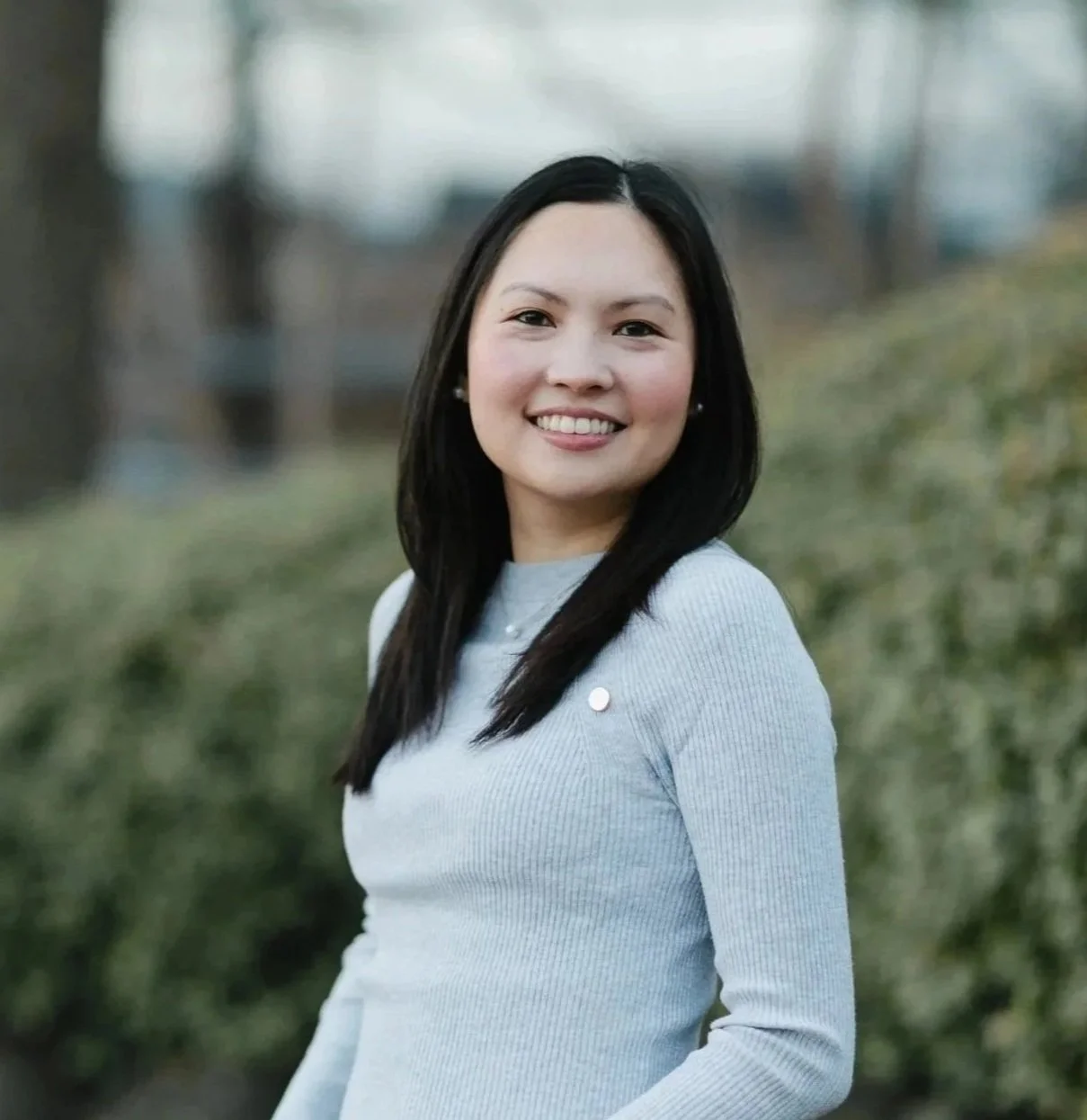 A young woman with long black hair and a light gray sweater smiling outdoors with blurred trees and greenery in the background.