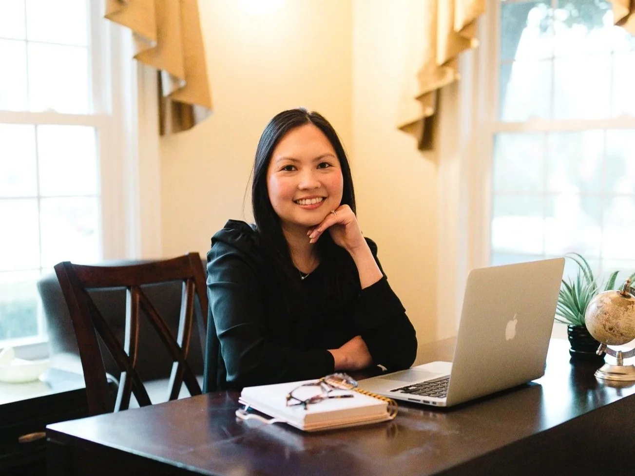 A woman sitting at a wooden desk with a laptop, glasses, a notebook, and a globe, smiling at the camera in a well-lit room with large windows.