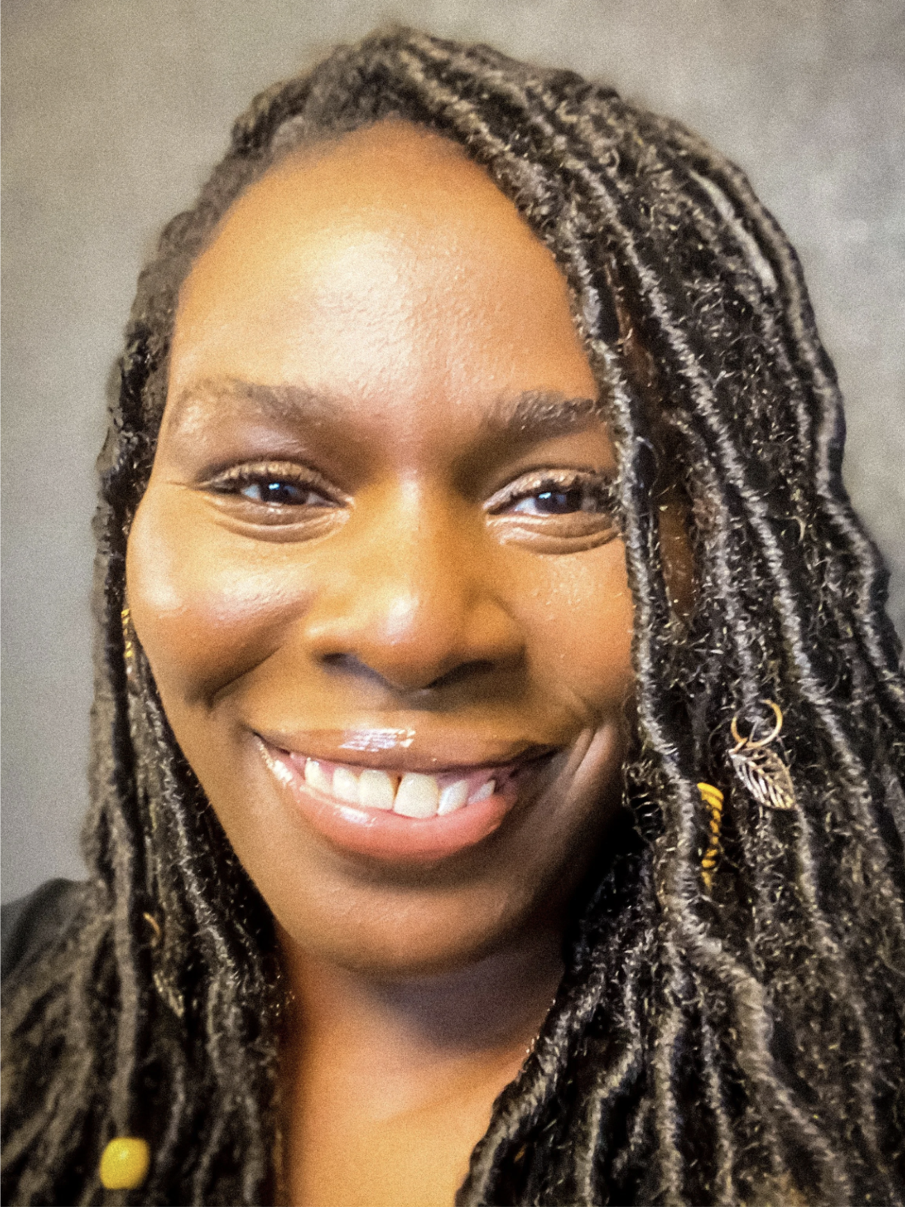 A close-up of a young African American woman smiling, showing white teeth, with braided hair decorated with gold beads and charms, and wearing earrings.