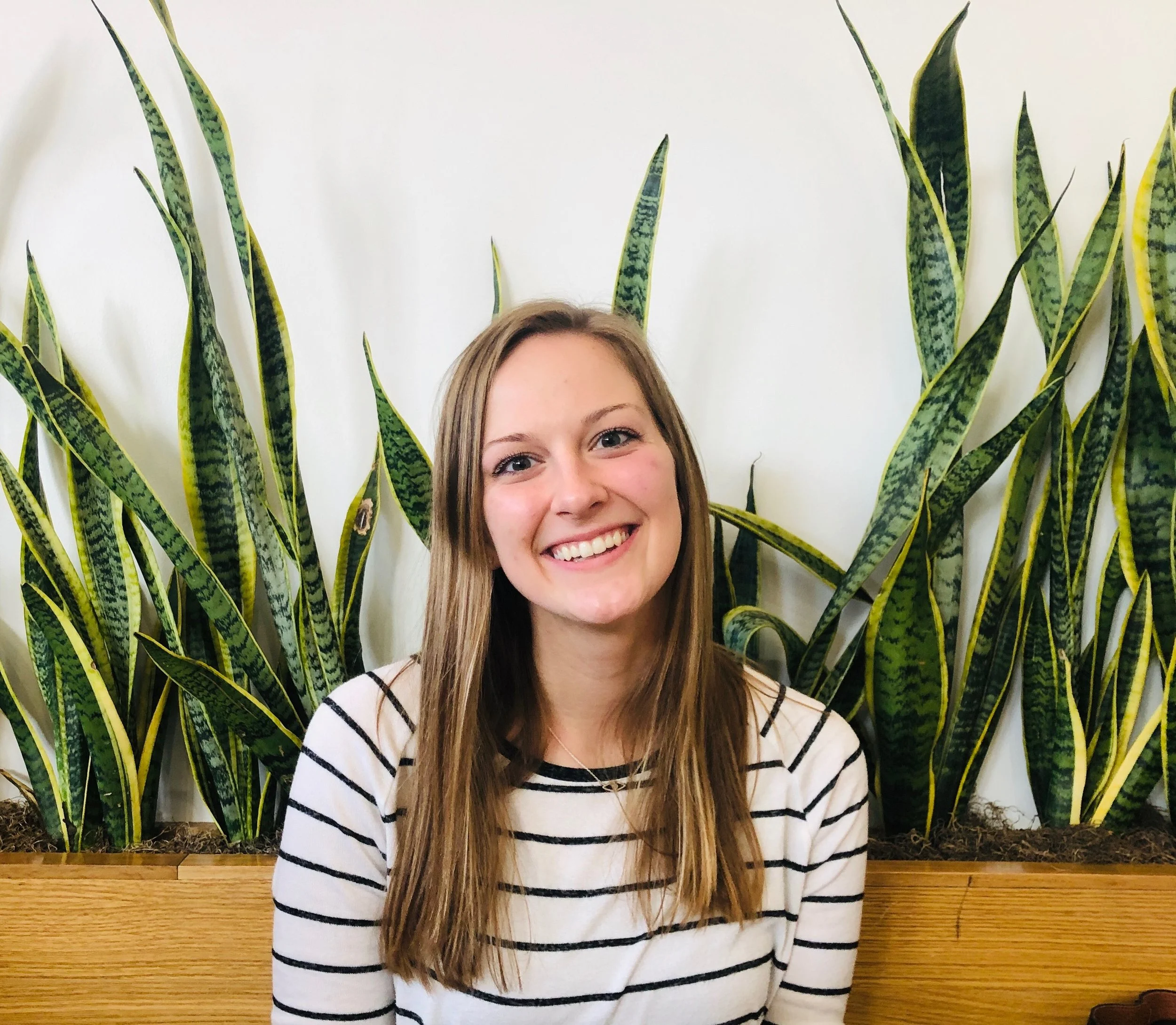 Young woman with long blonde hair and a striped shirt smiling, standing in front of tall green plants with a white background